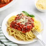 A plate of spaghetti topped with rich Italian meat sauce and shredded cheese is served with a fork on the side. A sprig of basil garnishes the dish alongside savory meatballs. In the background, there is a bowl of grated cheese and another dish with more sauce. A green and white striped napkin is placed nearby.