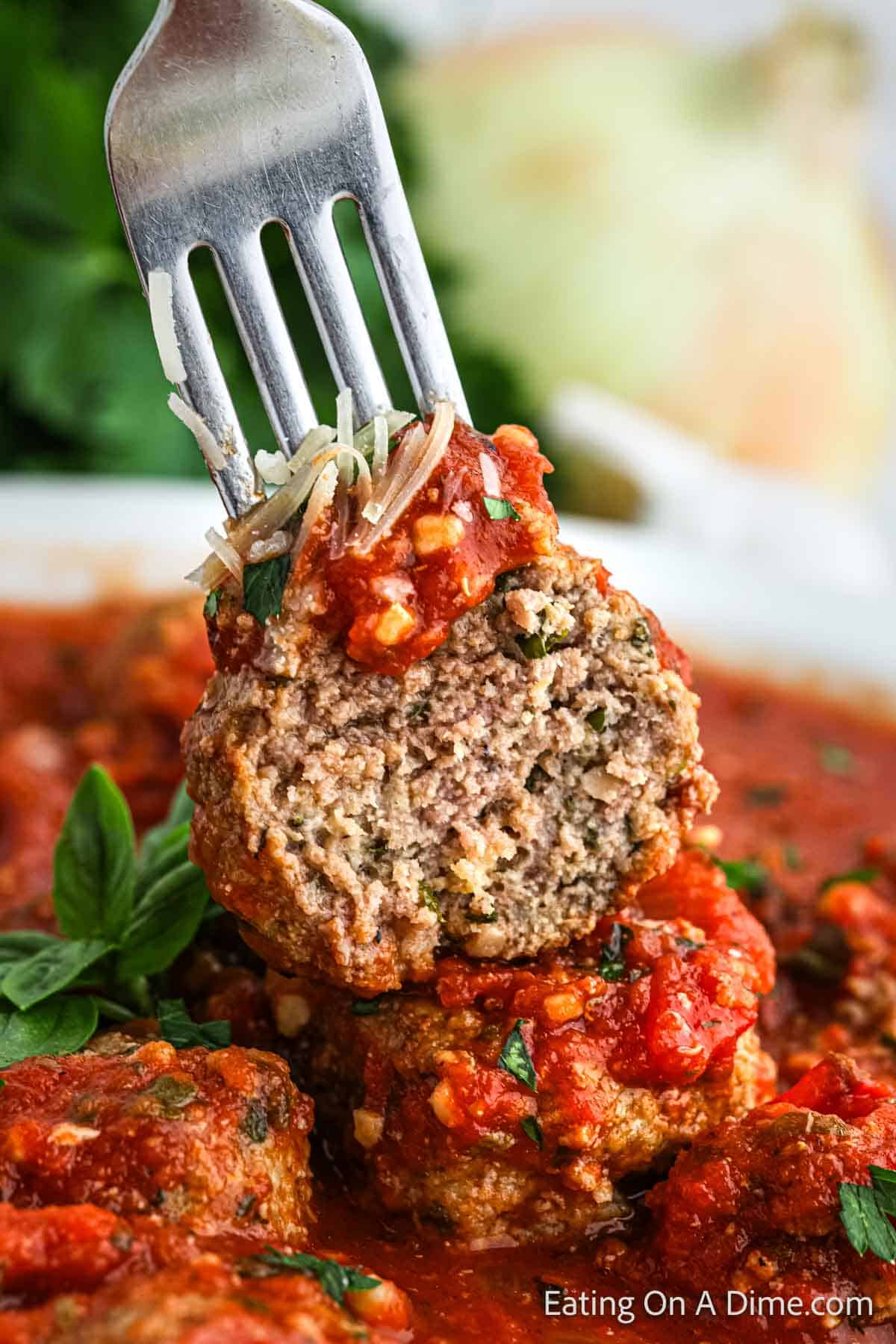 A close-up of a fork holding an Italian meatball covered in chunky tomato sauce, showing a bite revealing the inside texture. Fresh herbs and additional Italian meatballs with tomato sauce are visible in the background.