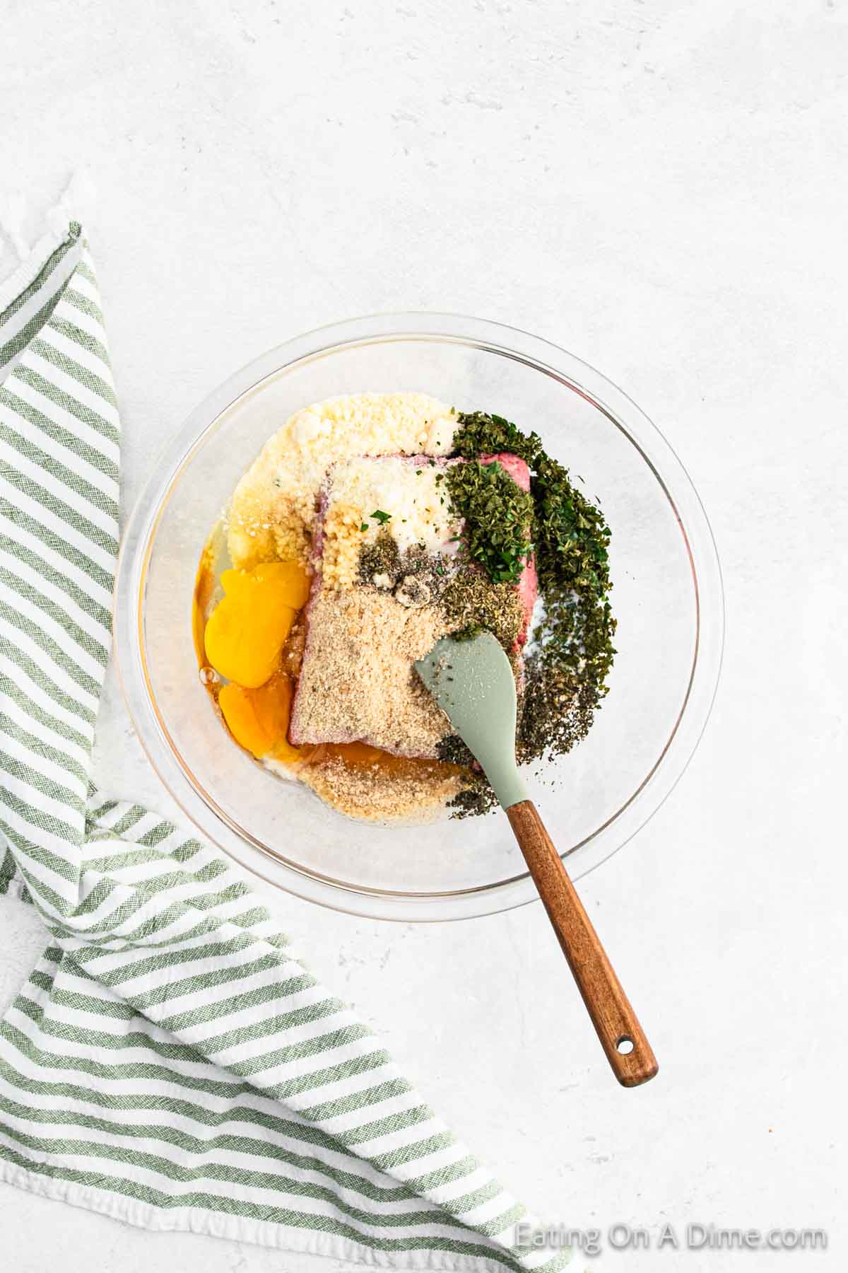 A clear glass bowl contains raw eggs, breadcrumbs, chopped herbs, and seasoning for Italian meatballs. A green spatula with a wooden handle is resting inside the bowl. A green and white striped kitchen towel is placed on the left side of the bowl on a white countertop.