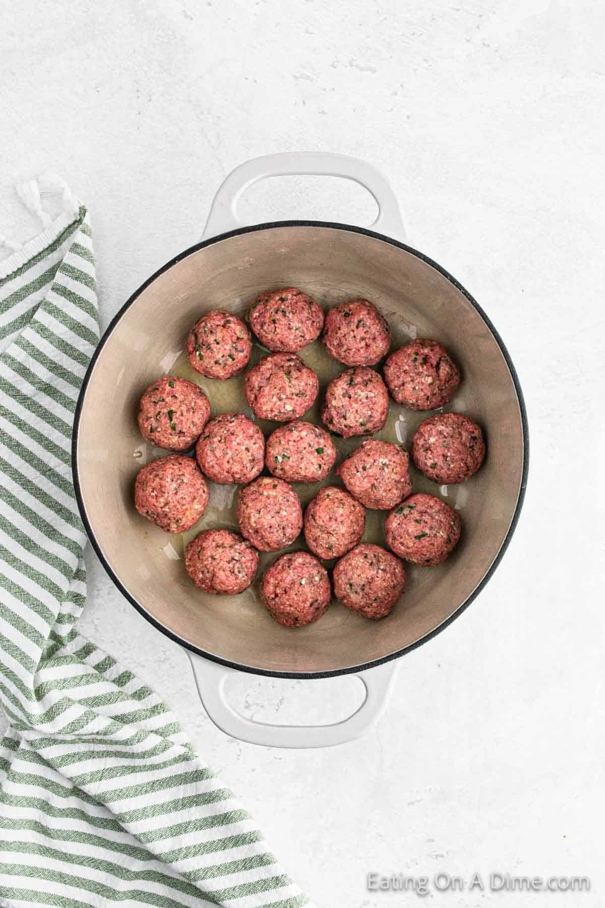 A round, white Dutch oven containing uncooked meatballs arranged in a single layer on the bottom. The Dutch oven rests on a light-colored surface next to a folded green and white striped kitchen towel, ready to start an Italian recipe.