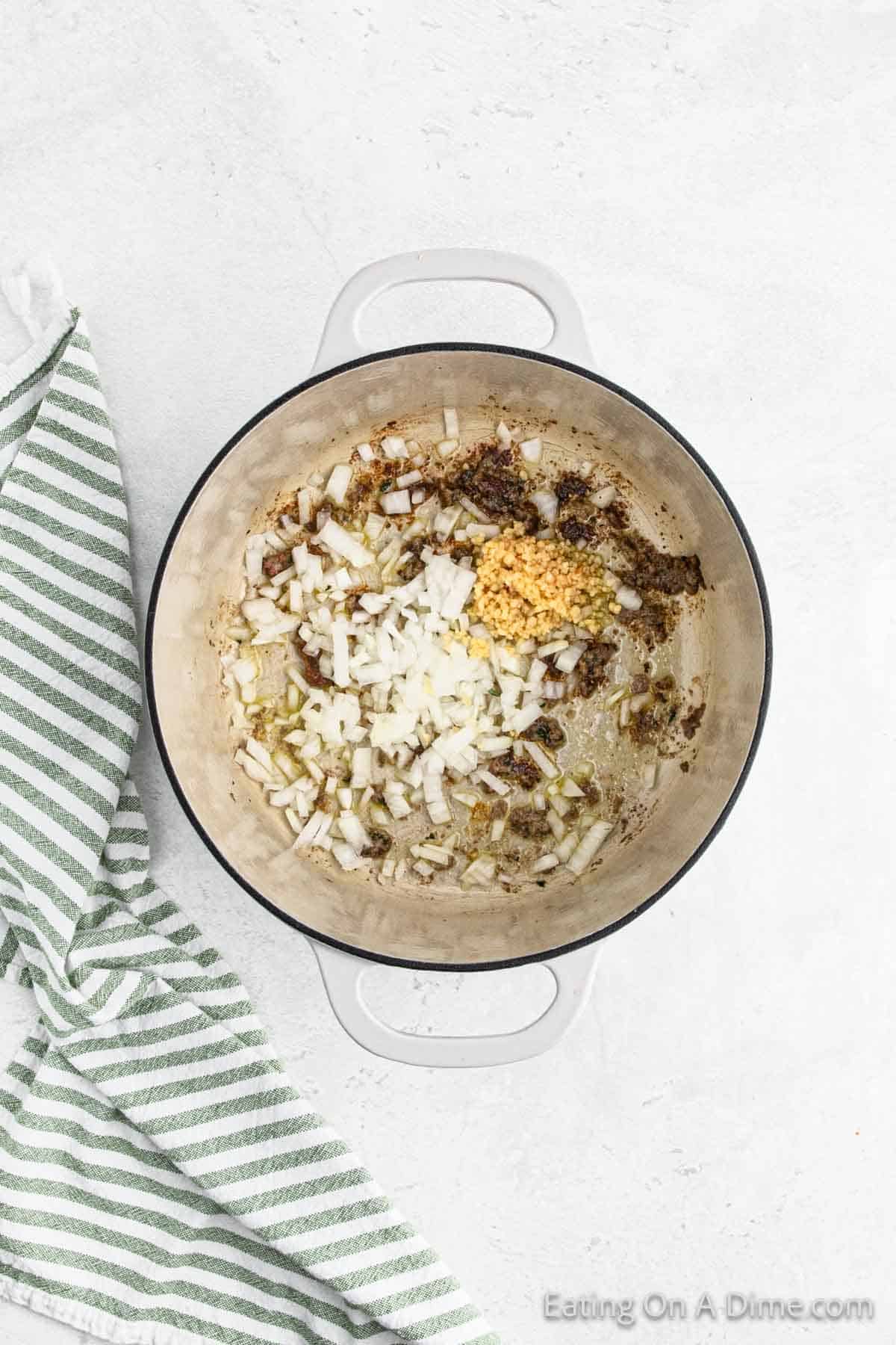 A large white pot on a light-colored surface, containing chopped onions and minced garlic cooking in oil, sets the stage for an Italian recipe. A green and white striped kitchen towel is placed to the left of the pot, ready for the addition of savory meatballs.