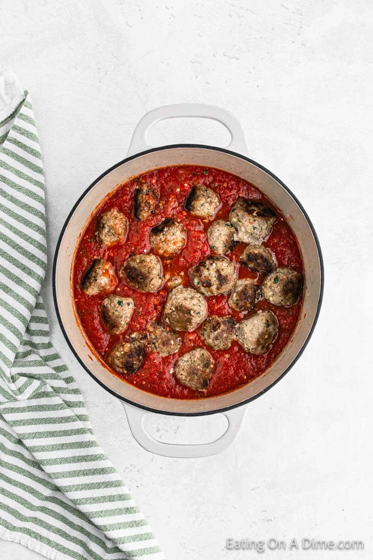 A white ceramic pot filled with Italian meatballs simmering in red tomato sauce sits on a light gray surface. A green and white striped kitchen towel is partially visible next to the pot on the left side.