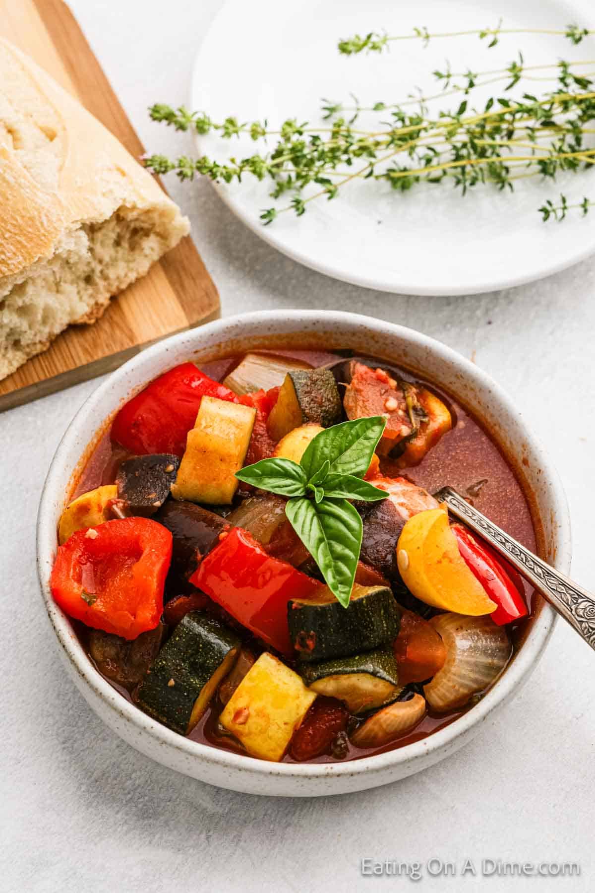 A bowl of cooked zucchini, squash, onion and tomatoes in a bowl of sauce with a spoon with a side of crusty bread. 