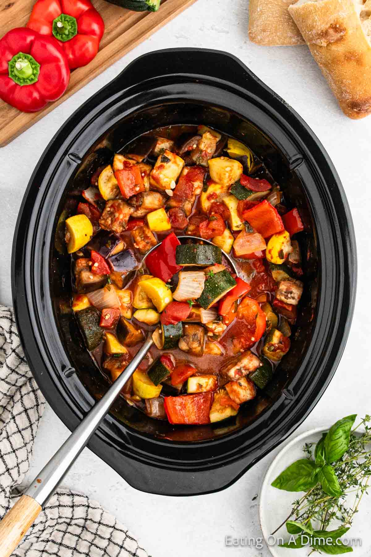 Yellow squash, zucchini, egg plant and red bell pepper cooked in a tomato based sauce in the slow cooker with a silver ladle. 