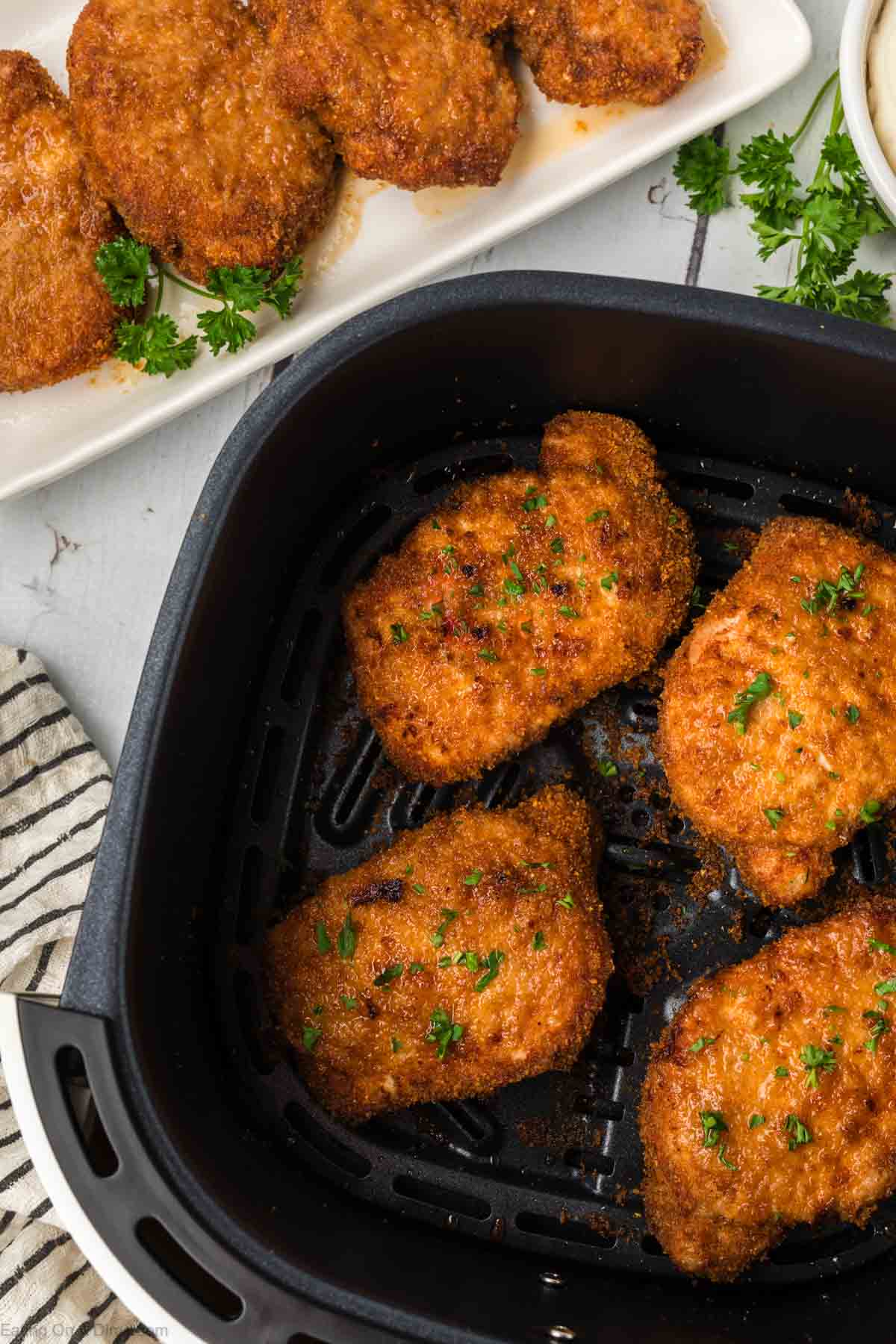 Shake and Bake Pork Chops in the air fryer basket with a 4 shake and bake pork chops on a platter on the side