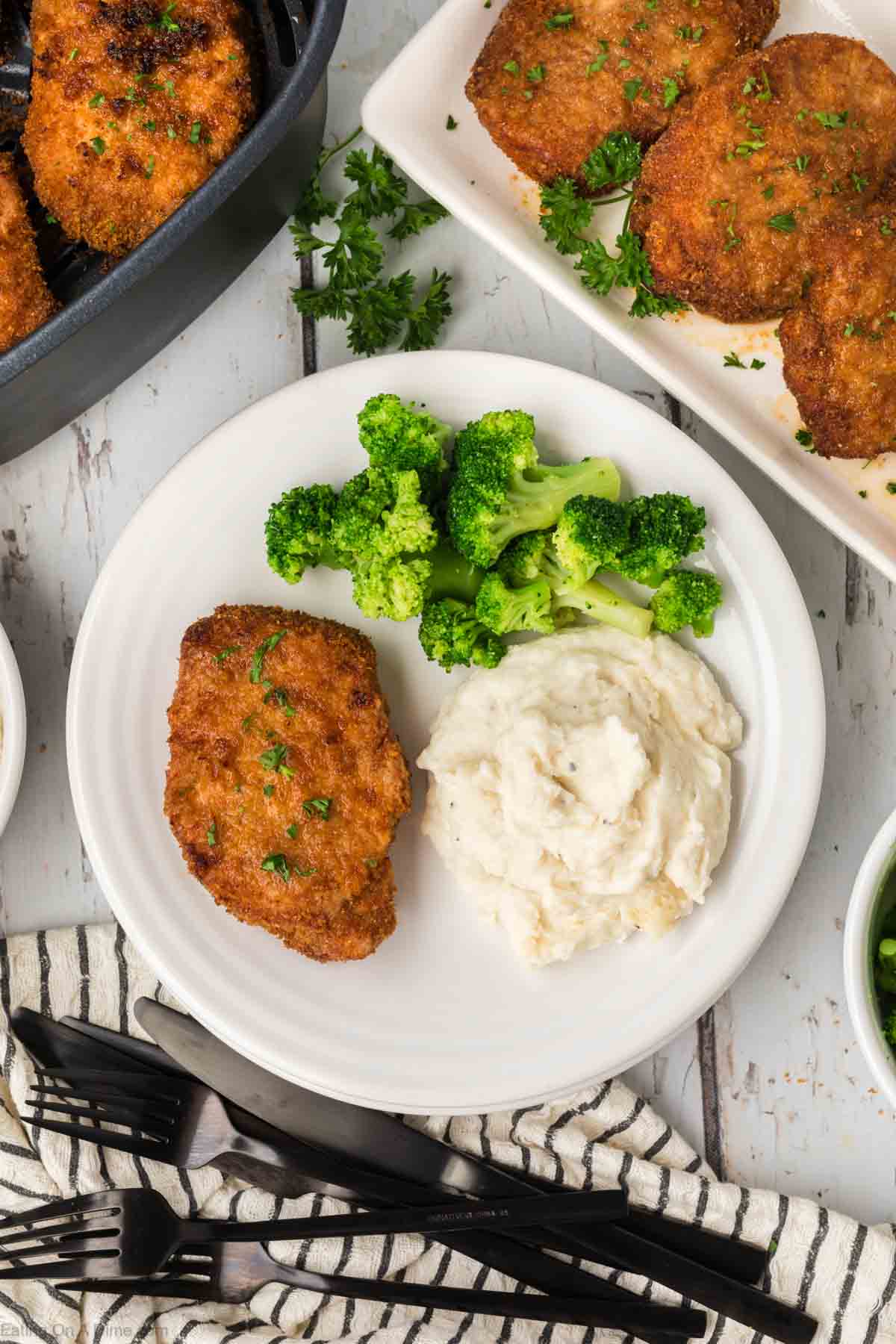 Shake and bake pork chops on a plate with mashed potatoes and broccoli on the side with a platter of pork chops on the side