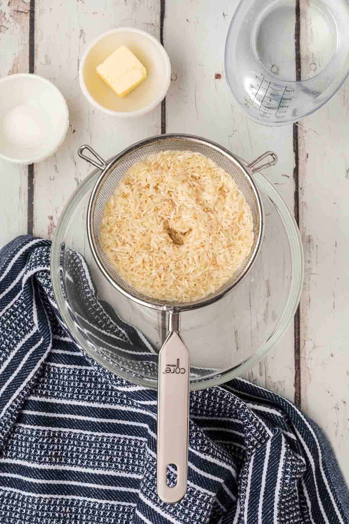 Rinsing uncooked rice in a strainer over a bowl with small bowls of salt, butter and water