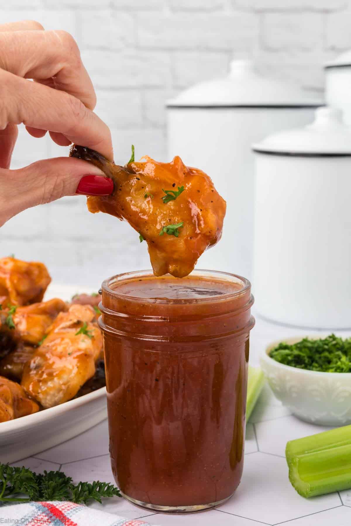 Wing sauce in a jar with a chicken wing covered in sauce is over the top with a platter of chicken wings on the side