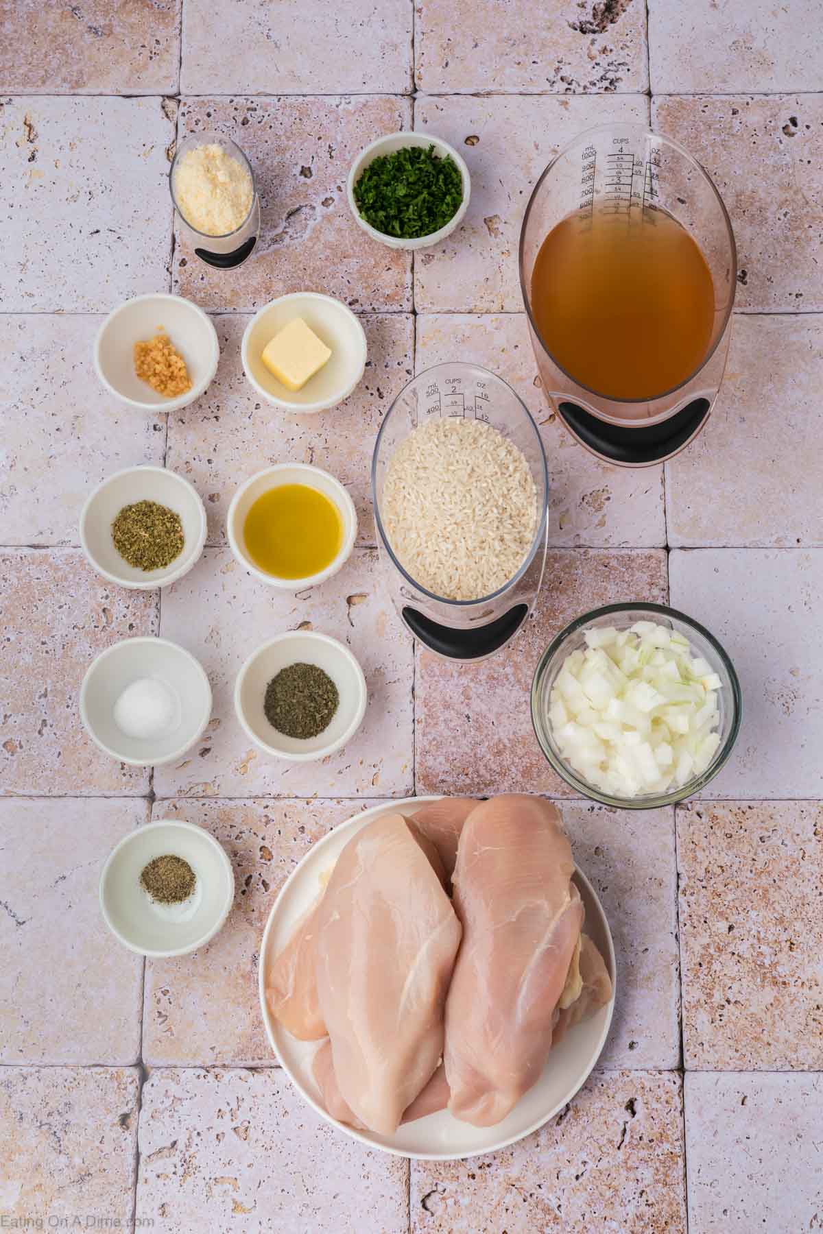 An assortment of ingredients on a tiled surface including raw chicken breasts on a plate, rice, chopped onions, olive oil, butter, grated cheese, minced garlic, chicken broth, and various seasonings in small bowls.