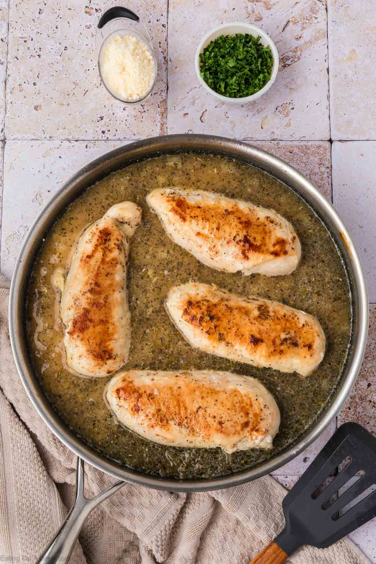 A skillet with four browned chicken breasts simmering in a savory sauce. A spatula rests on the handle of the skillet. In the background, there's a small bowl of grated cheese and another filled with chopped herbs, all set on a textured white surface, ready to be served over fluffy rice.