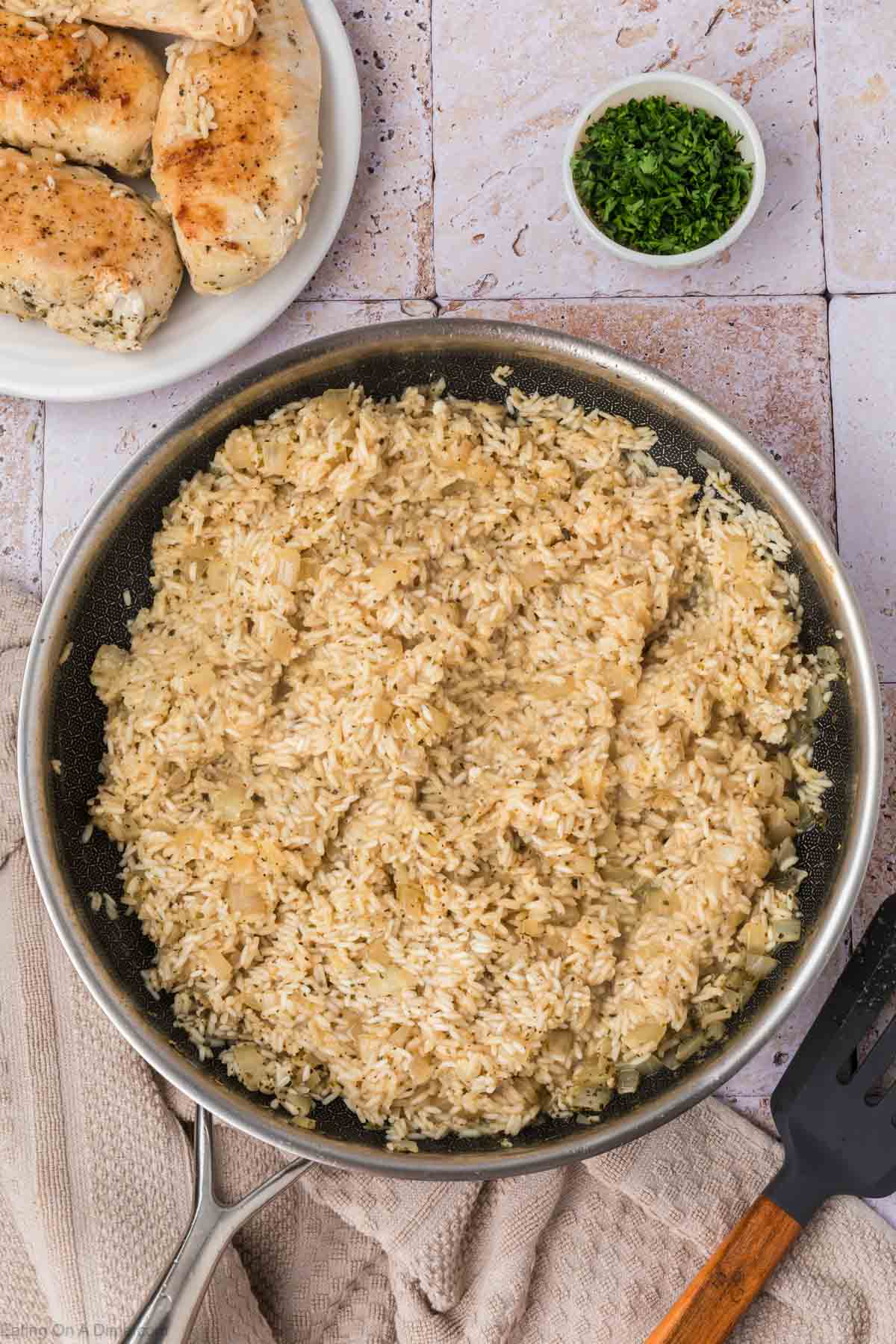 A skillet filled with seasoned rice is placed on a light-colored tiled surface. To the top left, a plate holds two pieces of cooked, seasoned chicken. A small bowl of chopped green herbs is above the skillet. A black spatula and beige towel are nearby.