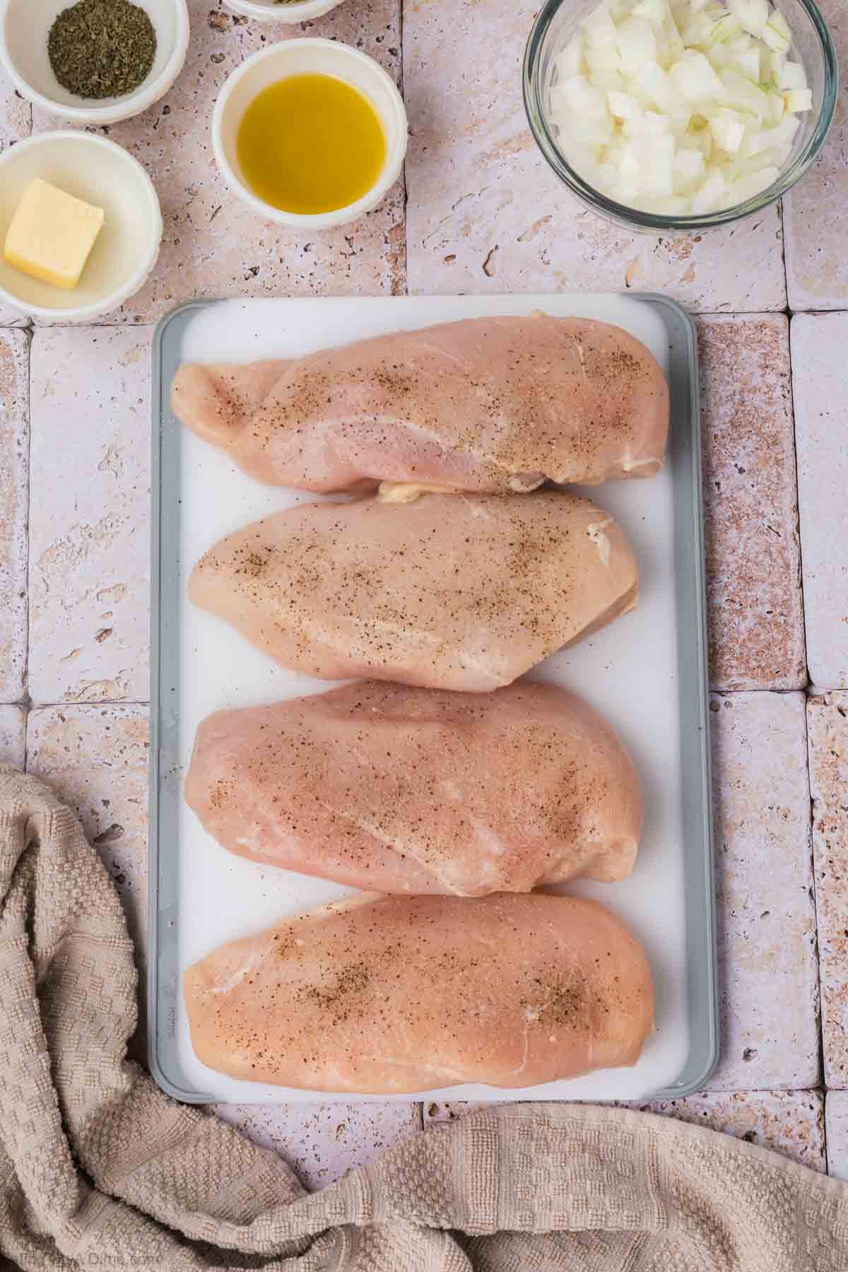 An overhead shot of four raw chicken breasts seasoned with salt and pepper on a white cutting board. Surrounding the board are small bowls containing olive oil, chopped onions, butter, and herbs. A textured cloth is partially visible in the bottom left corner, alongside a bowl of uncooked rice.