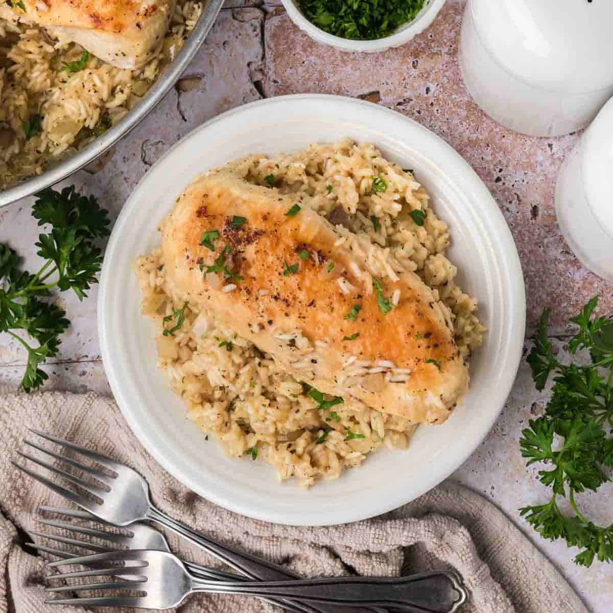 A white plate with a serving of seasoned chicken breast placed on a bed of creamy rice garnished with fresh parsley. The dish is surrounded by chicken pieces, a neutral-colored textured cloth, a bundle of forks, and white condiment containers on a rustic surface.