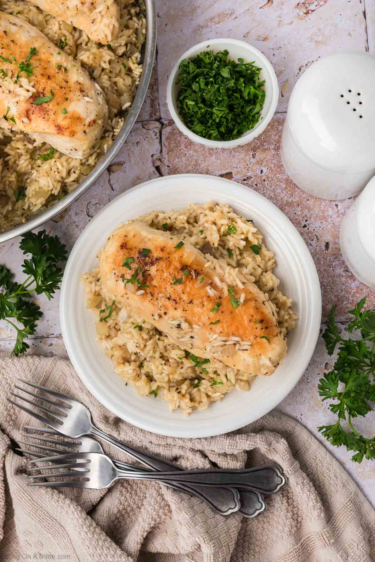 A plate of baked chicken served on a bed of seasoned rice, garnished with chopped parsley. The plate is placed on a rustic surface next to a bowl of more parsley, salt and pepper shakers, a skillet brimming with additional chicken and rice, and a folded beige cloth with two forks.