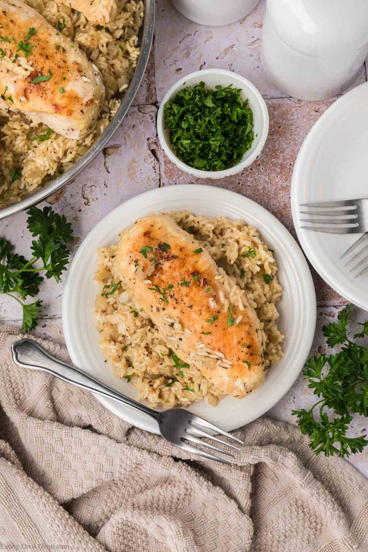 A plate with a serving of golden-brown chicken breast over a bed of creamy rice, garnished with herbs. A bowl of chopped greens, a pan with more chicken and rice, a beige napkin, and white plates with forks are visible around the table.