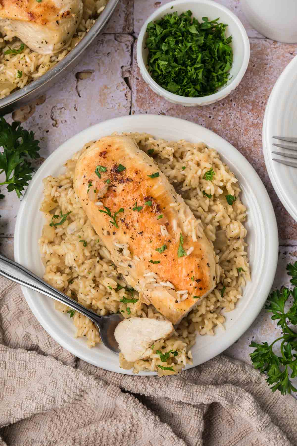 A white plate is filled with seasoned grilled chicken breast on a bed of fluffy rice, garnished with chopped parsley. A fork, holding a tender piece of chicken, rests on the side. In the background, there’s a small bowl of chopped parsley and part of a white dish towel peeking into view.