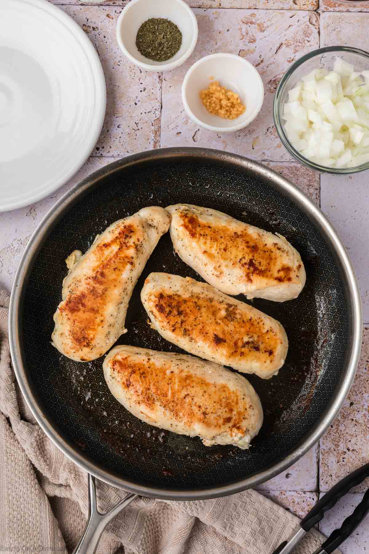 Four browned chicken breasts are in a frying pan. Surrounding the pan are small bowls containing chopped onions, minced garlic, dried herbs, and an empty white plate. A light brown towel is partially visible under the pan. The background is a tiled surface, ready for a delicious Chicken and Rice dish.