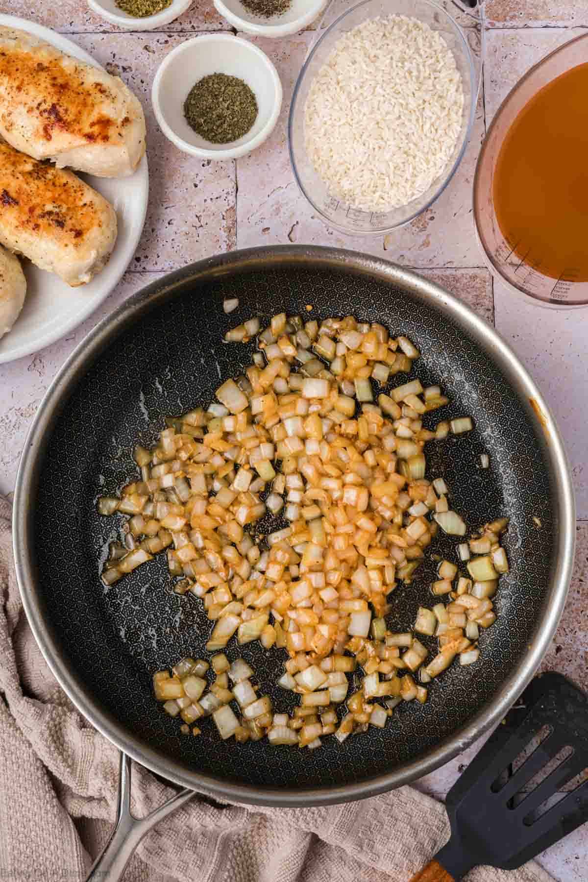 A frying pan with diced onions sautéing in it is placed on a kitchen counter. Surrounding the pan are a plate with cooked chicken breasts, bowls containing seasonings and uncooked rice, and a measuring cup with broth. A black spatula lies next to the pan, ready to assist in your chicken and rice culinary creation.