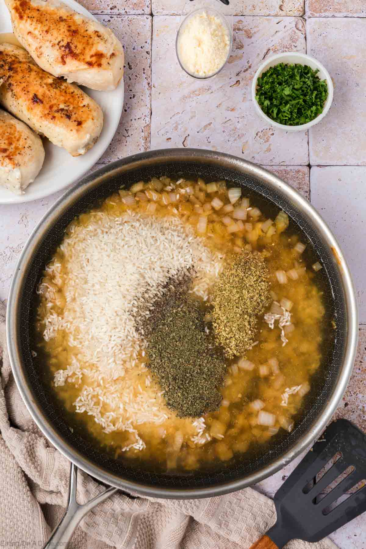 A top-down view of a skillet filled with broth, rice, chopped onions, and various seasonings, ready to be cooked. In the background, there's a plate with cooked chicken breasts and small bowls containing grated cheese and chopped parsley. A spatula and a beige cloth are nearby—perfect for Chicken and Rice.