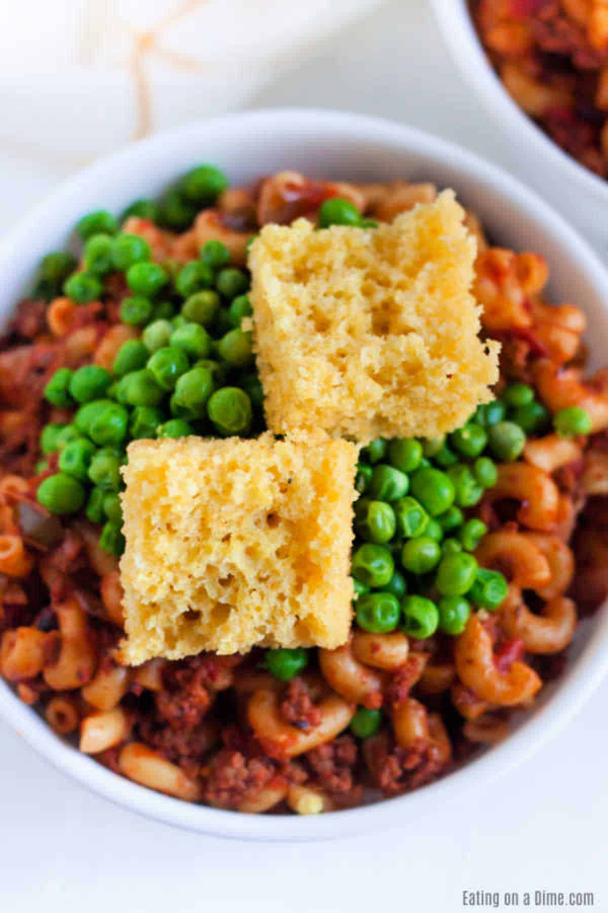Goulash in a bowl with macaroni noodles with a tomato base meat mixture topped with peas and corn bread