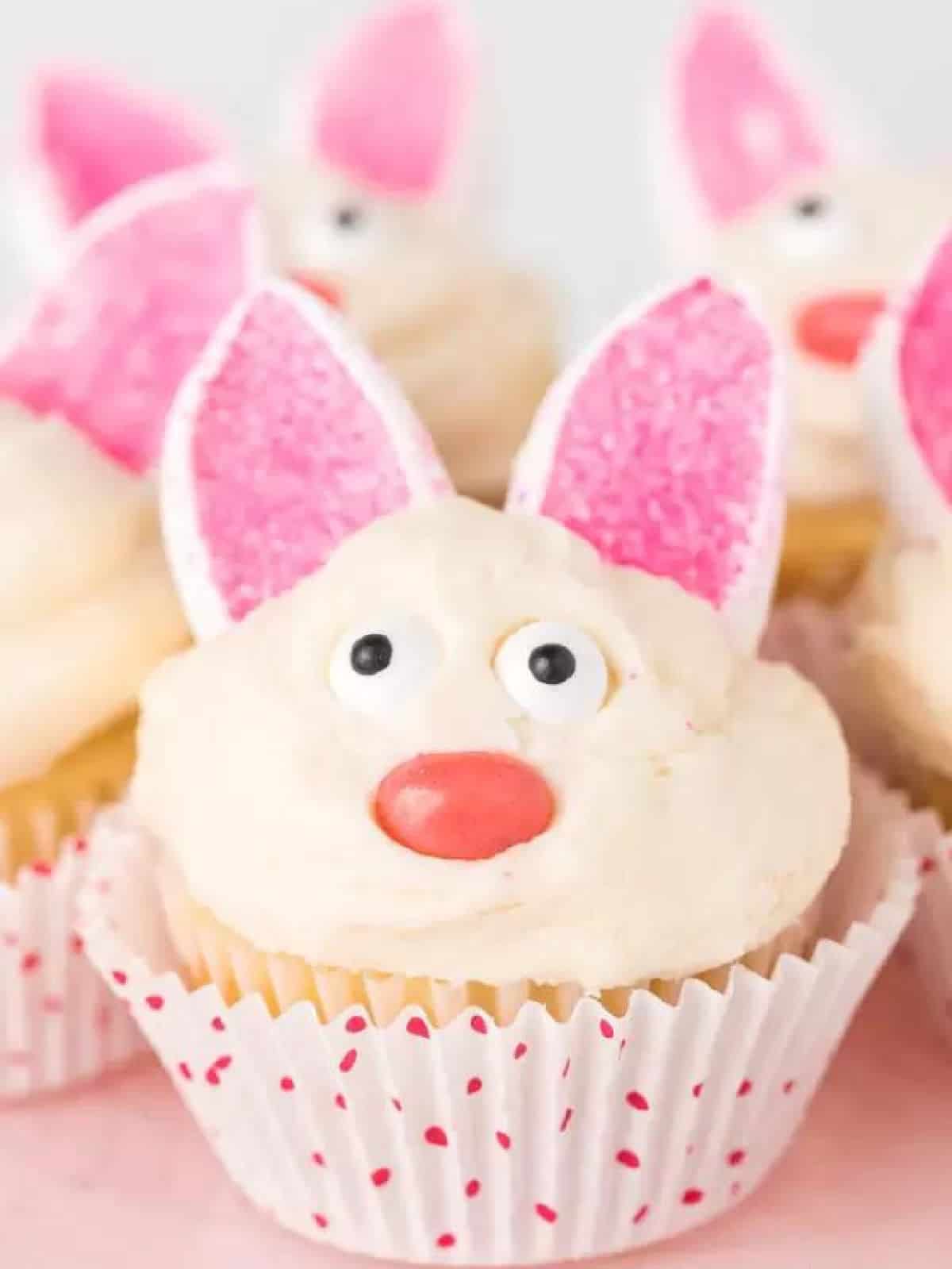 Easter Bunny Cupcakes with pink sprinkle ears with candy eyes and a jelly bean nose with white frosting in a cupcake liner with poke a dots