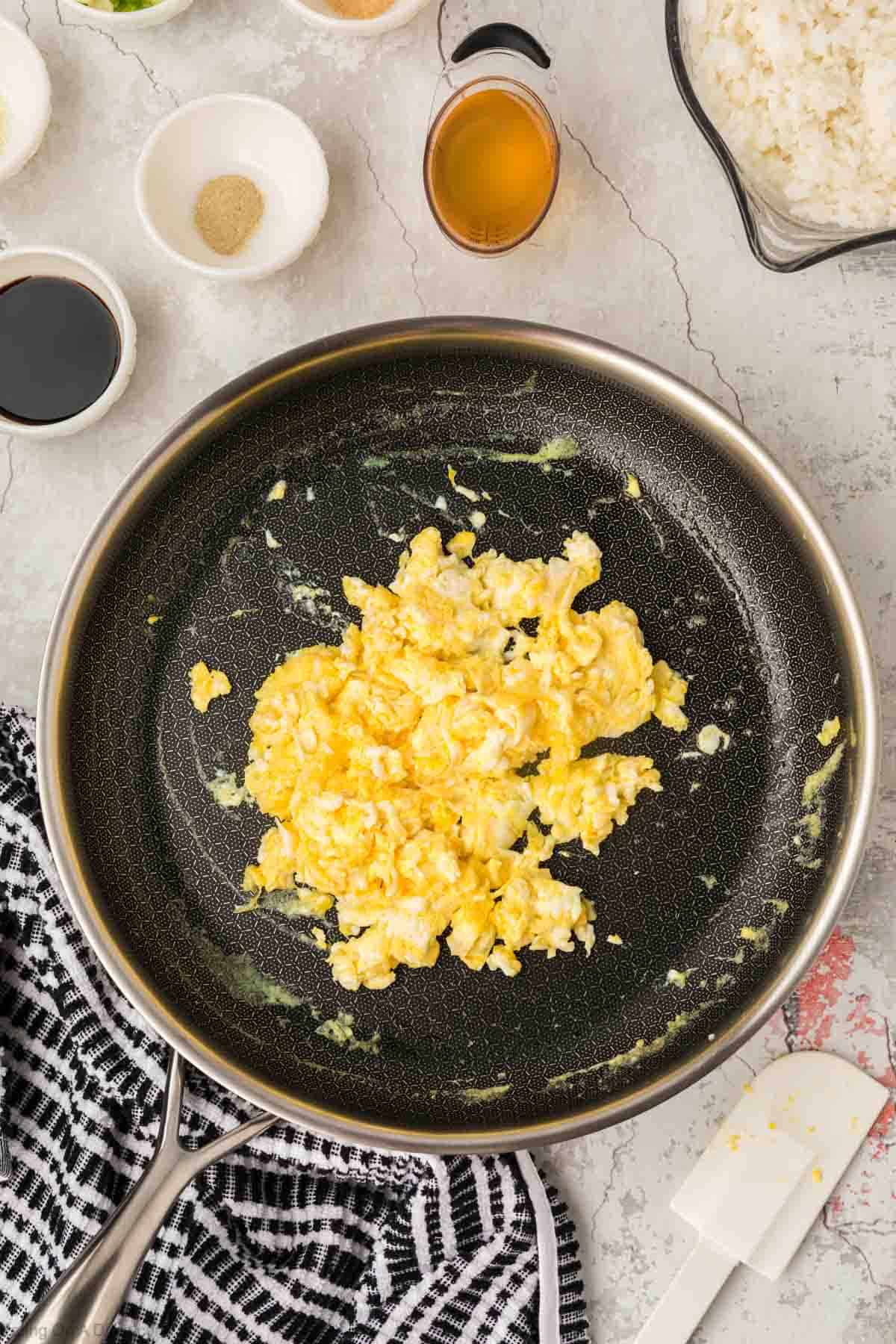 Scrambled eggs in a skillet with small bowls of soy sauce, garlic powder, and rice