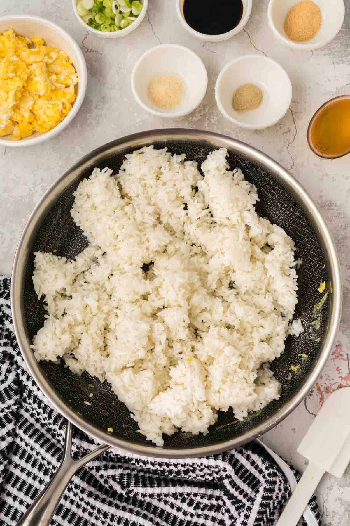 Rice in a skillet with a bowl of scrambled eggs in a bowl, green onions, garlic powder, onion powder, and soy sauce