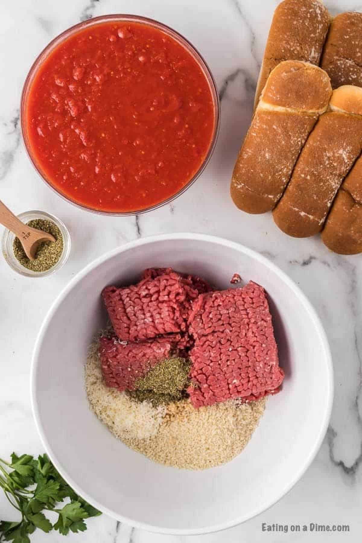 Raw ground beef in a bowl with breadcrumbs, parmesan cheese, and seasoning with a bowl of sauce, seasoning and hoagies