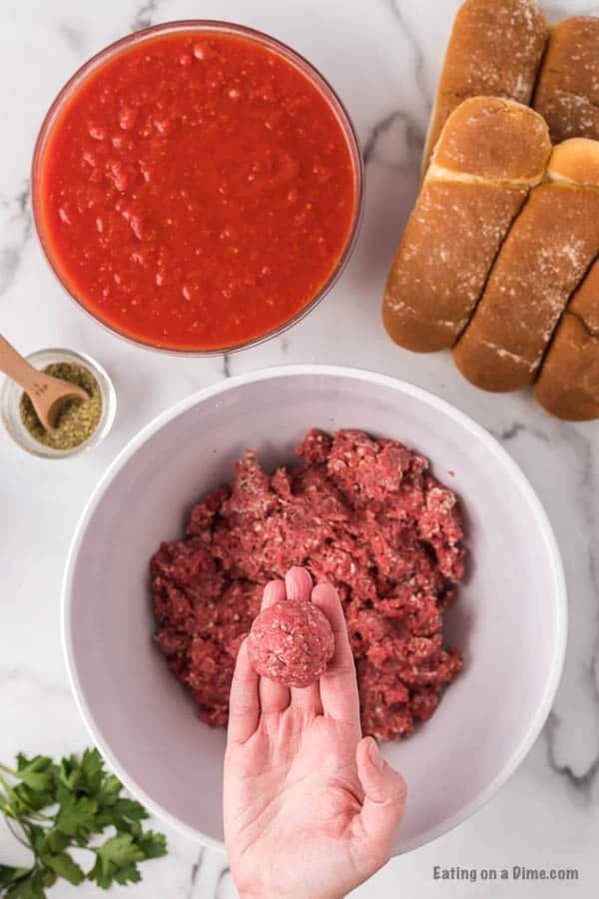 Ground beef mixture in a bowl with a hand holding a meatball with a bowl of sauce on the side and seasoning