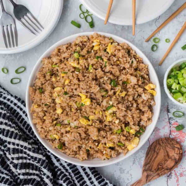 A bowl of egg fried rice with bits of scrambled egg and chopped green onions is placed on a marble surface. Surrounding the bowl are chopsticks, a small bowl of sliced green onions, a wooden spoon, a black and white kitchen towel, and two empty white plates with forks.