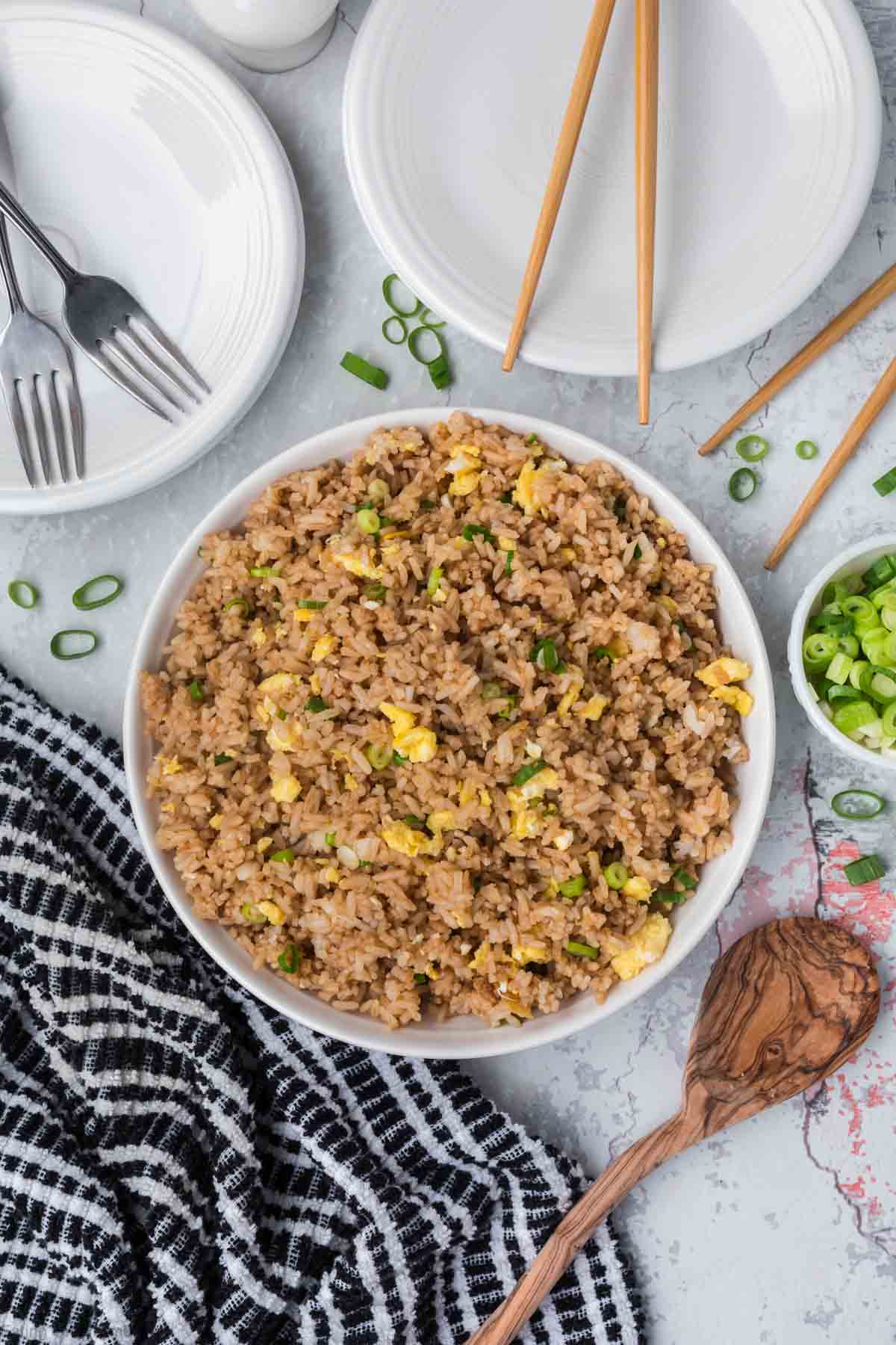 A bowl of easy egg fried rice with chopped scallions and scrambled eggs rests on a gray surface. Nearby are empty white plates with forks, chopsticks, and a wooden spoon. Beside the bowl, there's a small dish of sliced green onions and a black-and-white checkered napkin.