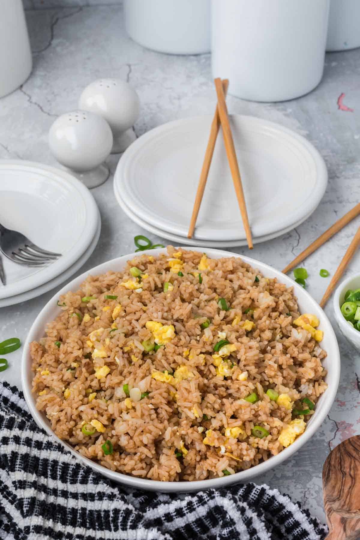 A white bowl filled with Egg Fried Rice, garnished with chopped green onions. The rice contains scrambled eggs and vegetables. Behind the bowl, there are empty white plates, chopsticks, a fork, and a black and white checkered cloth. Salt and pepper shakers are also visible.