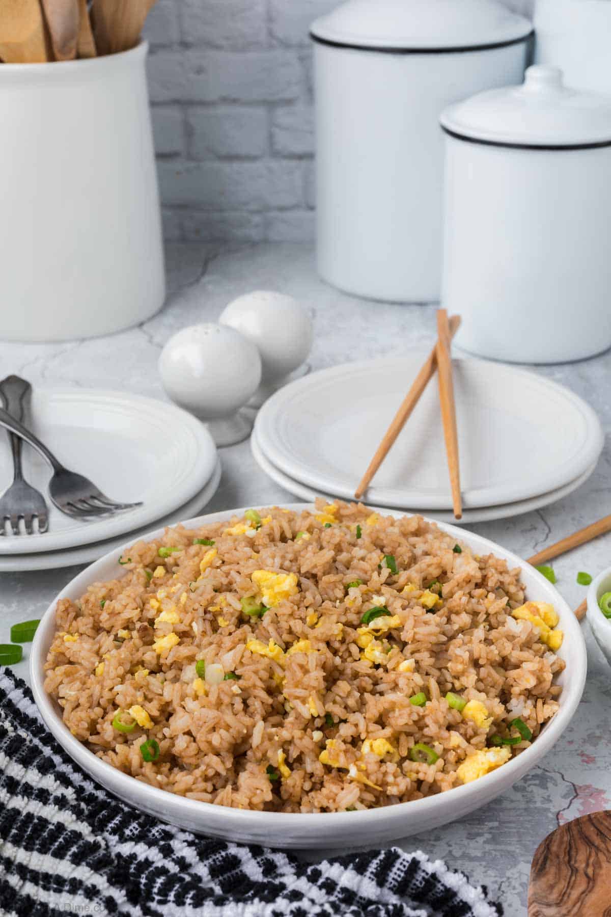 A bowl of Egg Fried Rice with scrambled eggs, green onions, and other ingredients is placed on a checkered cloth. In the background, there are stacked white plates with cutlery, chopsticks, white ceramic canisters, and two round salt and pepper shakers.