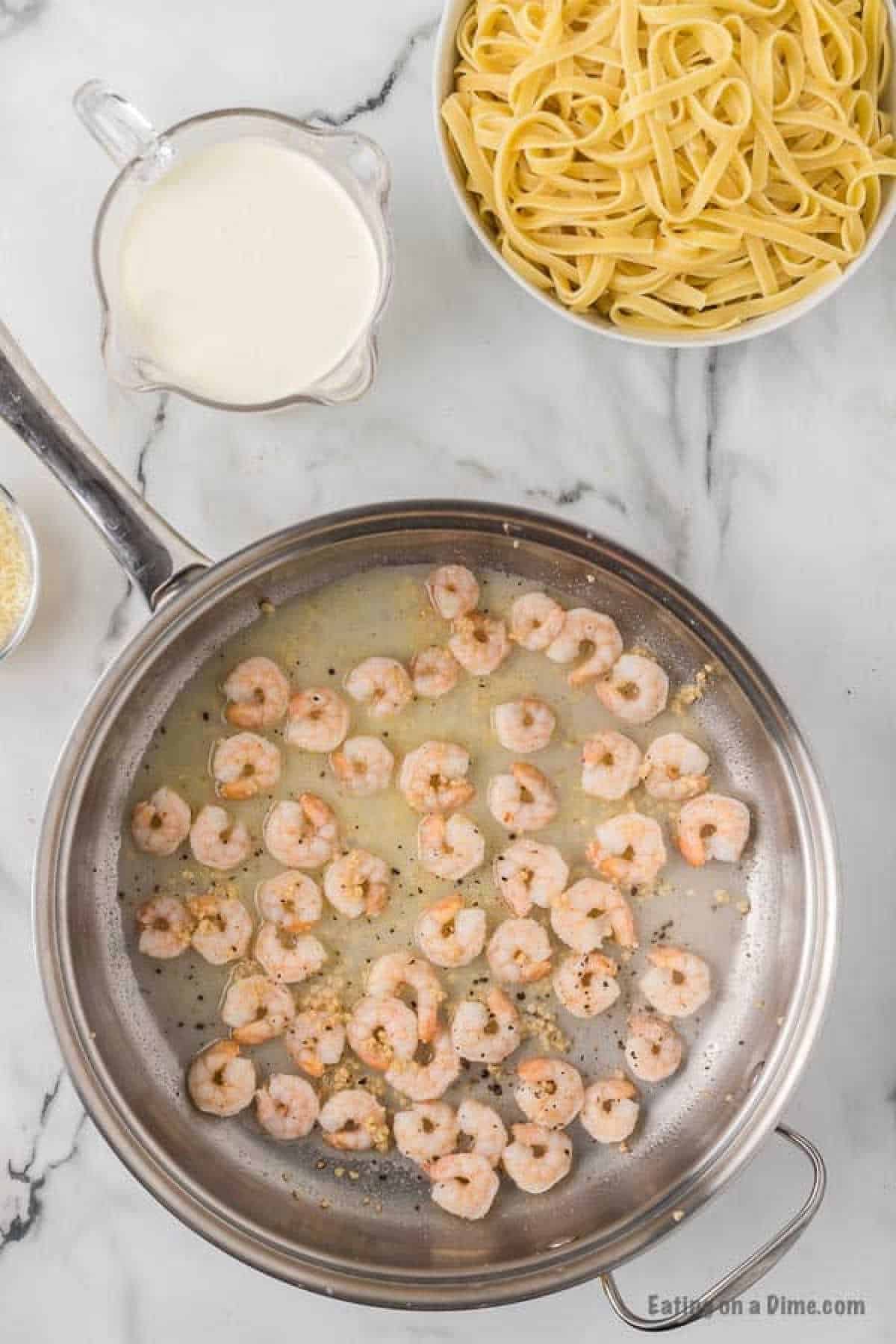 Cooking shrimp in a large skillet in a buttery sauce with seasoning. A bowl of fettuccini noodles and cream on the side