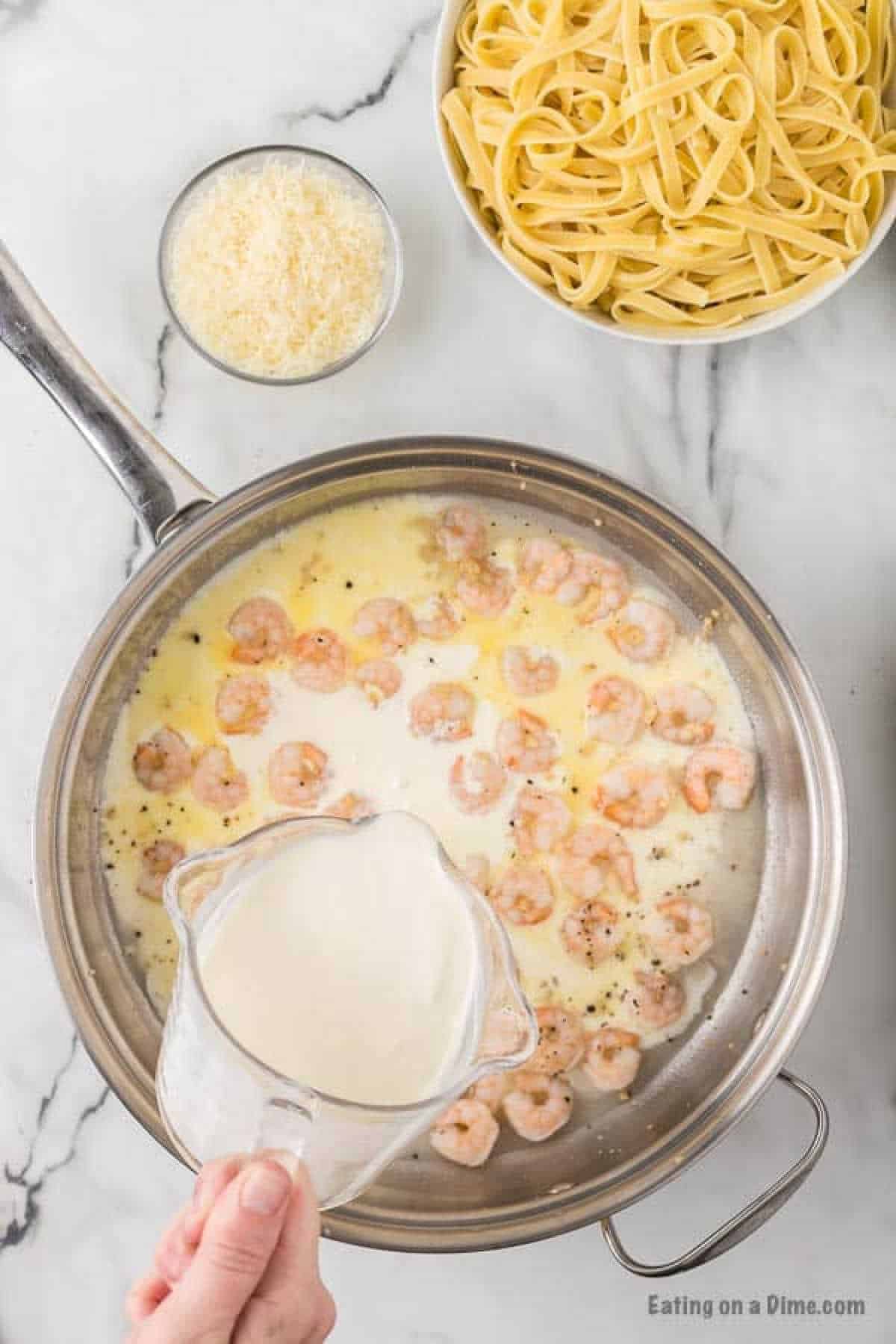 Pouring the cream in the skillet with the seasoned shrimp with a bowl of parmesan cheese and fettuccini noodles on the side