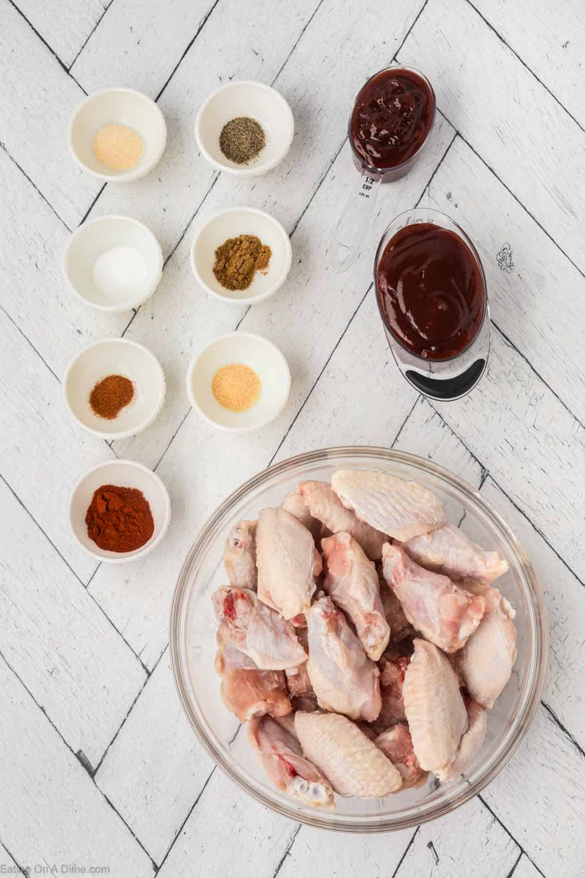 A flat lay image of ingredients for making BBQ chicken wings. There's a bowl of raw wings, small bowls containing various spices (black pepper, cumin, garlic powder, paprika, and chili powder), and two bowls with barbecue sauce. Perfect for a slow cooker meal!