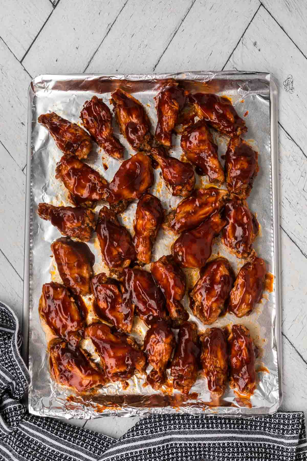 A baking tray covered with aluminum foil holds numerous golden-brown BBQ chicken wings glazed with a shiny barbecue sauce. The tray is set on a light gray wooden surface, with a dark blue and white striped cloth partially visible at the bottom left corner.