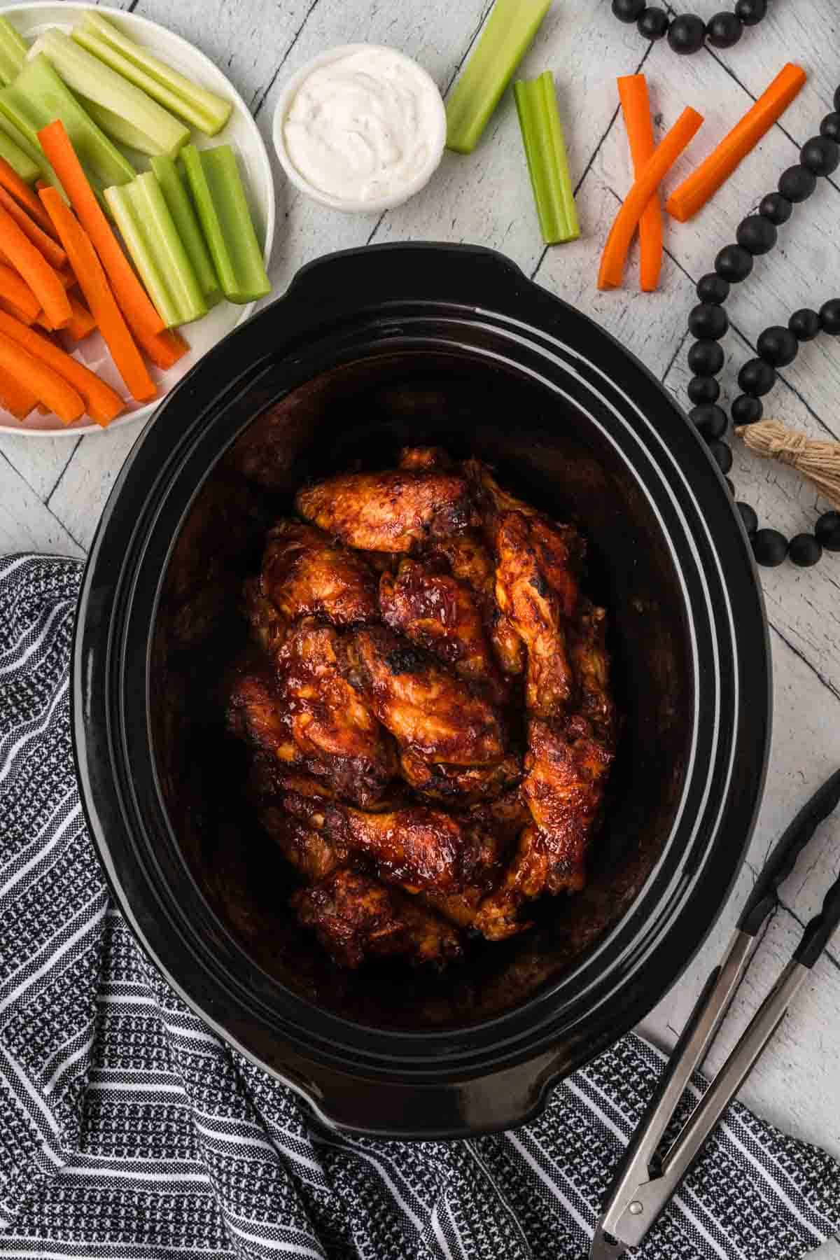 A black slow cooker filled with saucy BBQ chicken wings sits on a gray surface. Surrounding it are a plate of carrot and celery sticks with a small bowl of dip, beads, and kitchen tongs resting on a striped cloth.