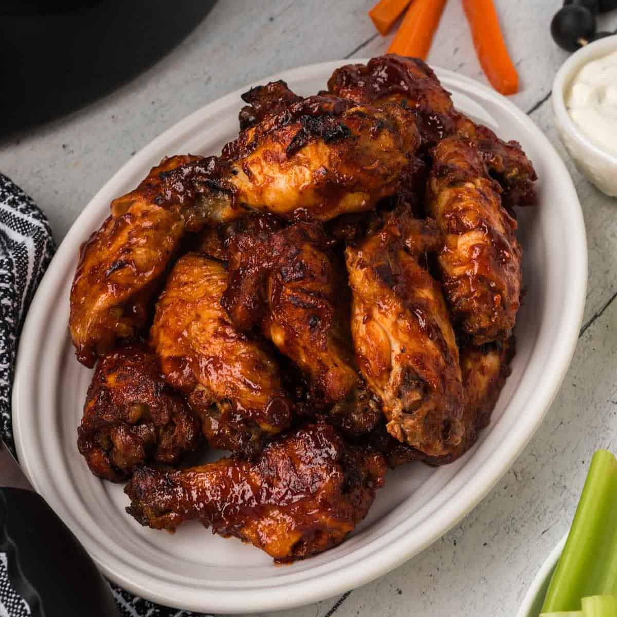 A white oval plate filled with glazed, crispy Slow Cooker BBQ chicken wings with a rich, dark sauce. The plate is set on a rustic white wooden surface, near a black and white napkin, a small bowl of ranch dressing, and some carrot and celery sticks in the background.