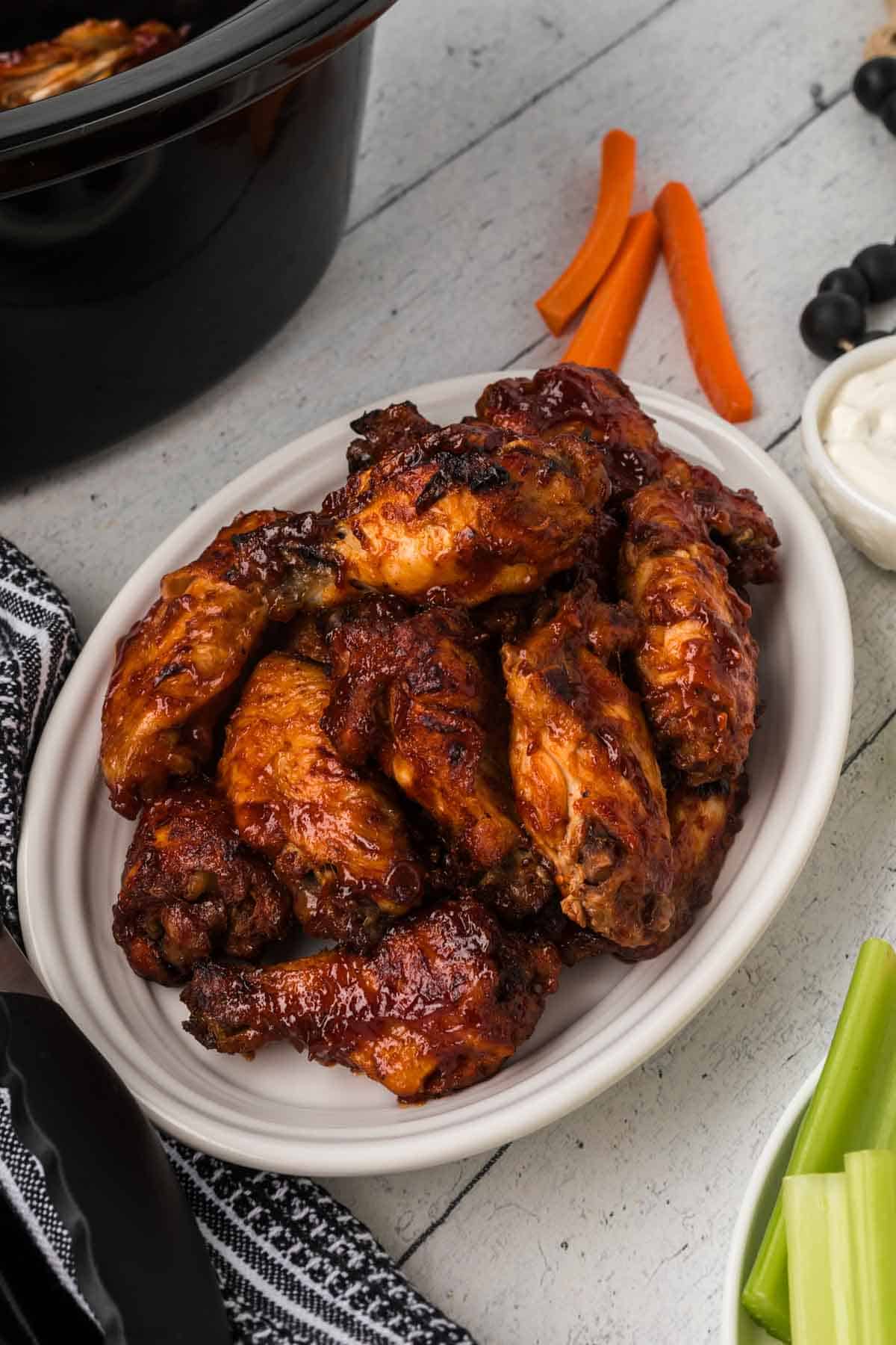 A white plate filled with slow cooker BBQ chicken wings coated in rich sauce sits next to carrot and celery sticks, a small bowl of white dipping sauce, and a black and white striped napkin on a rustic wooden table.