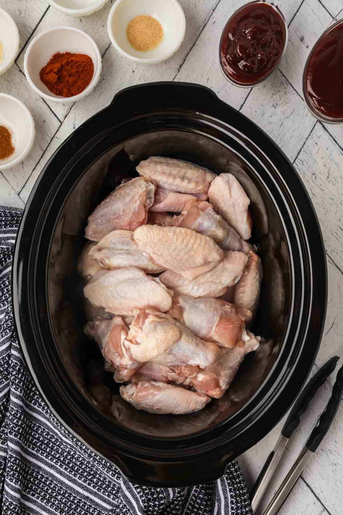 A black slow cooker filled with raw chicken wings is surrounded by small bowls containing various spices, seasonings, and BBQ sauces. A pair of tongs and a striped kitchen towel are placed nearby on a white wooden surface.