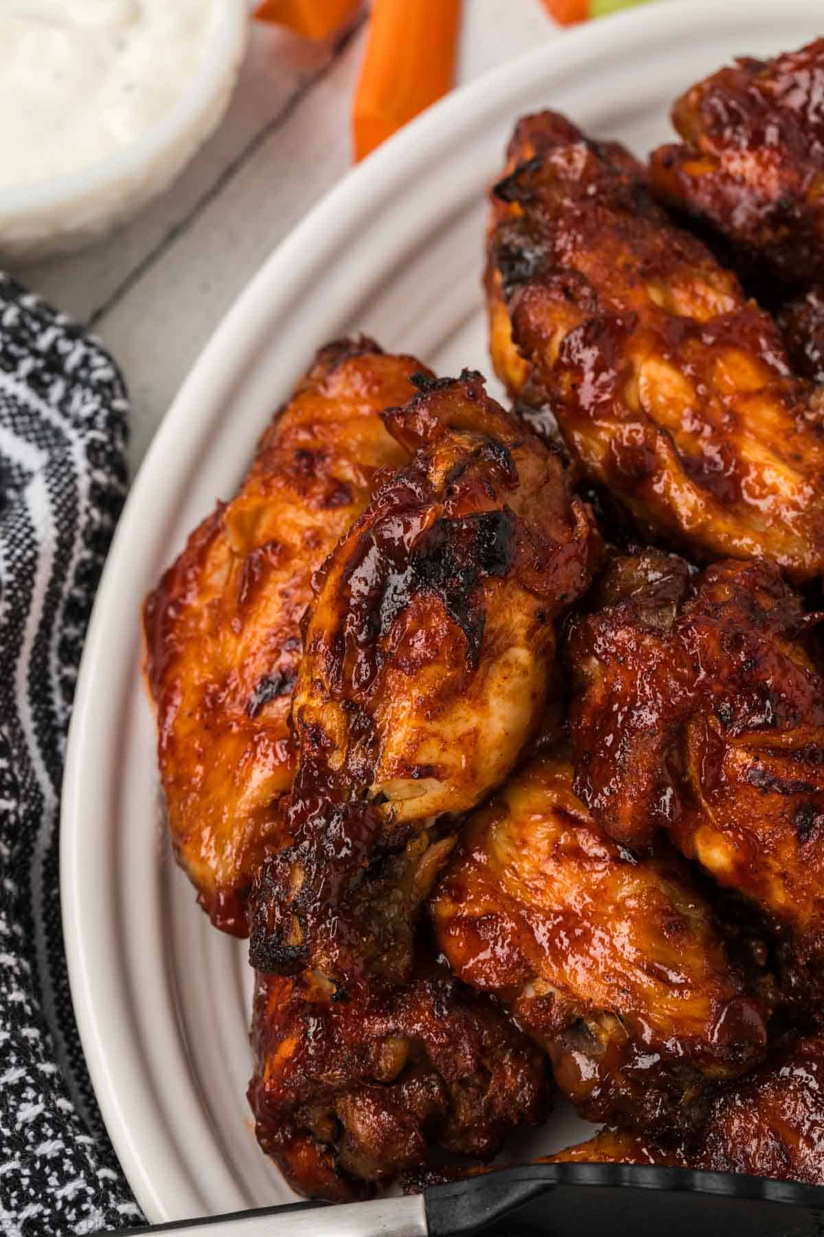 A close-up shot of a plate filled with crispy, glazed BBQ chicken wings. The wings are garnished with a caramelized, smoky sauce. Part of a black-and-white patterned cloth is visible on the left side of the plate, with a bowl of ranch dip and some carrot sticks in the background.