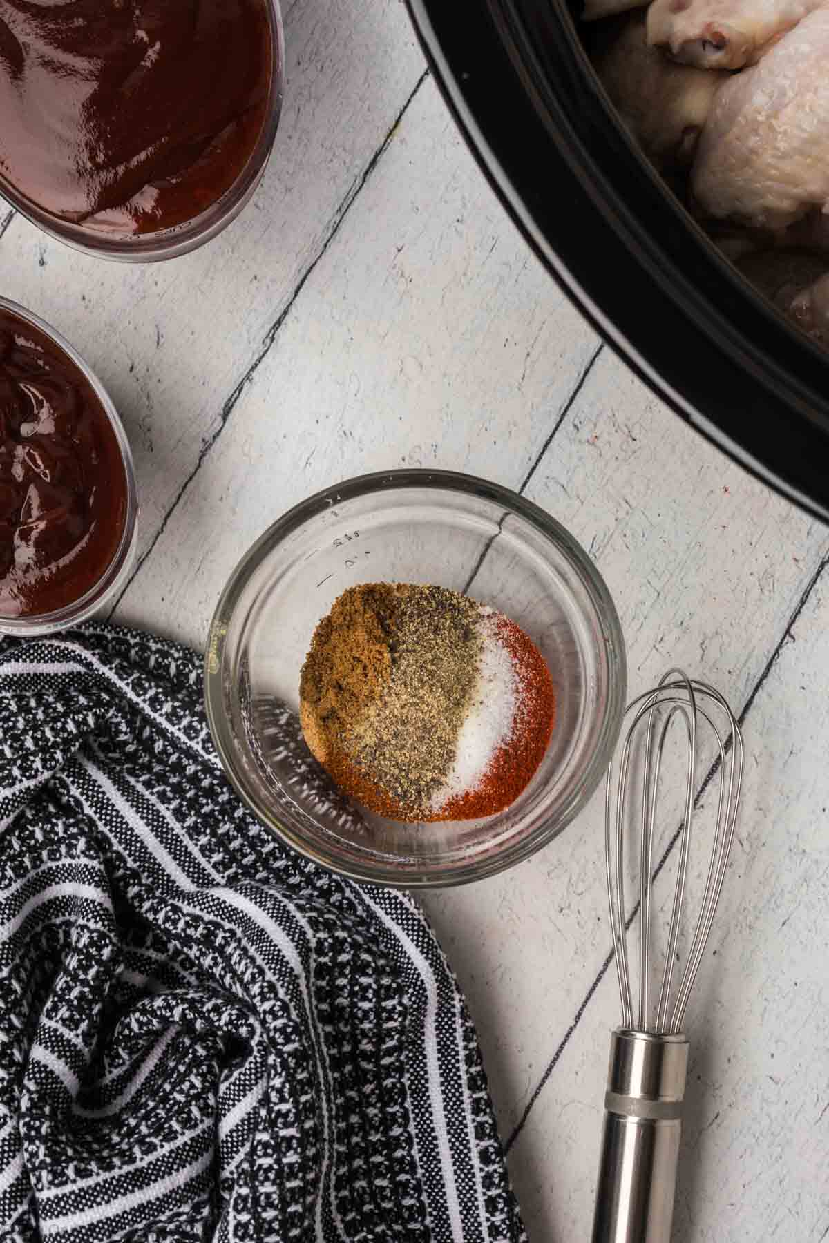 A small glass bowl containing a mixture of spices sits on a wooden surface next to a whisk and a black and white striped towel. Surrounding the bowl are jars of red sauce, and a slow cooker partially shown with BBQ chicken wings inside.