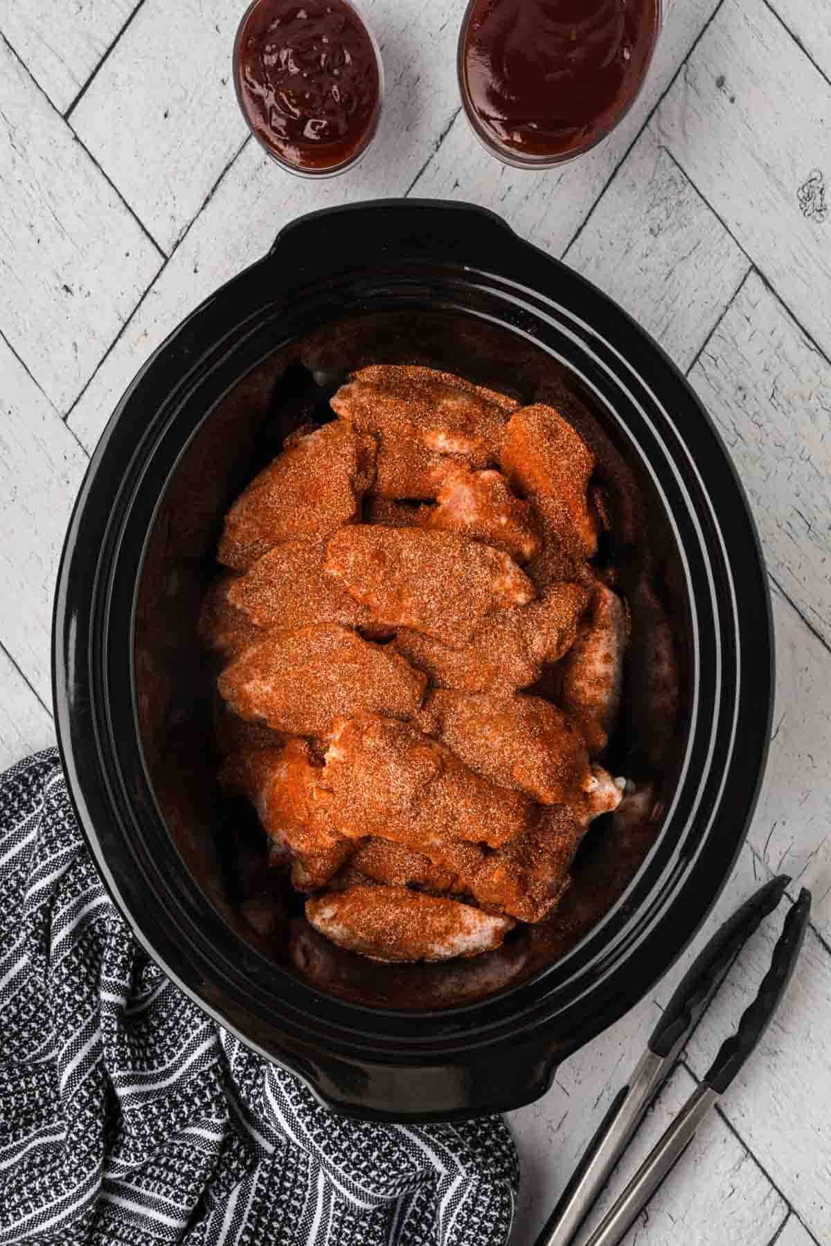 A black slow cooker filled with savory BBQ chicken wings rests on a light gray wooden surface. Beside it, two small bowls of sauces accompany a pair of metal tongs placed on a black and white patterned kitchen towel.