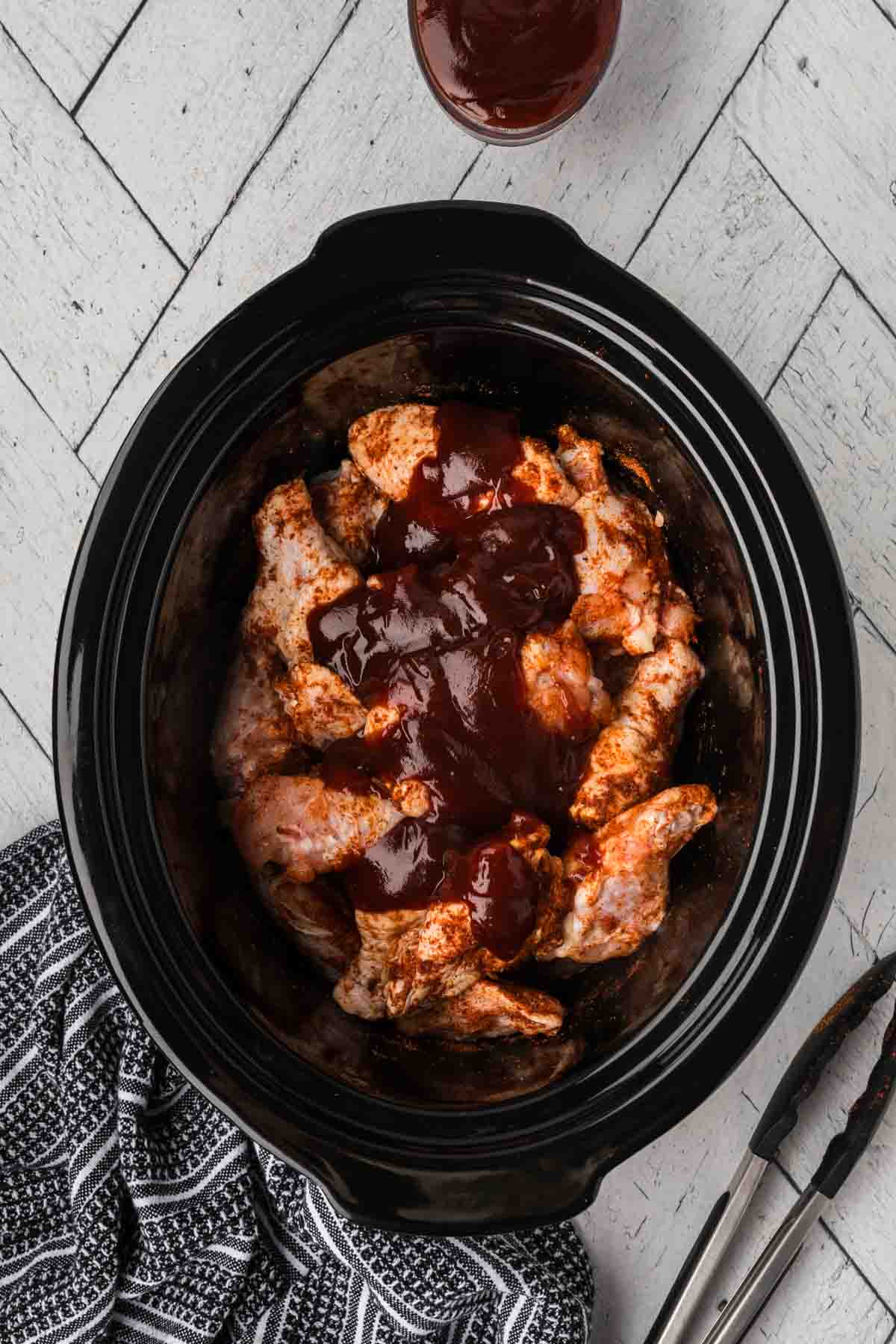 A slow cooker filled with BBQ chicken wings coated in barbecue sauce. Adjacent to the slow cooker are a pair of tongs and a black-and-white patterned cloth. A small container of barbecue sauce sits in the top right corner on a textured white surface.