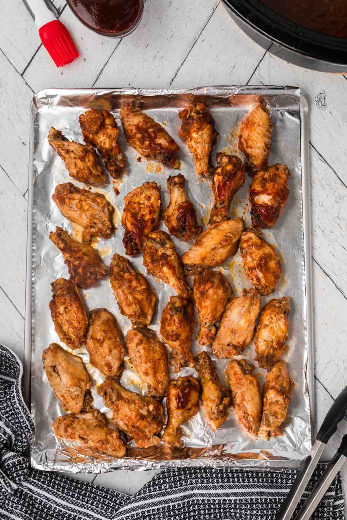 A baking sheet lined with foil is filled with a single layer of BBQ chicken wings, some slightly coated in sauce. A black and white striped cloth is at the bottom left corner, and a pair of tongs rests on the right side. A bowl of sauce with a brush is at the top.