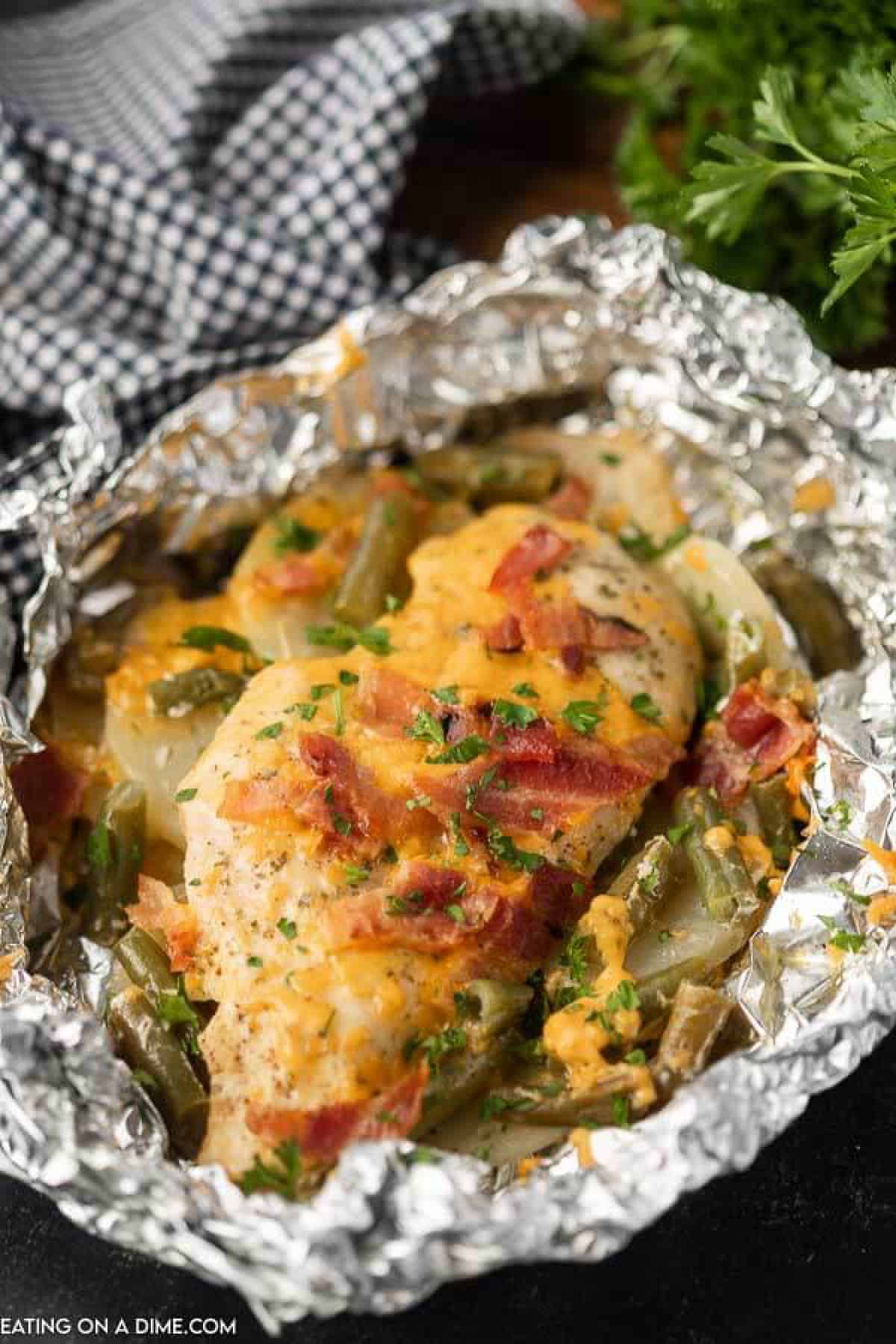 Cooked chicken breast in foil topped with diced bacon, melted cheese green beans and slice potatoes 