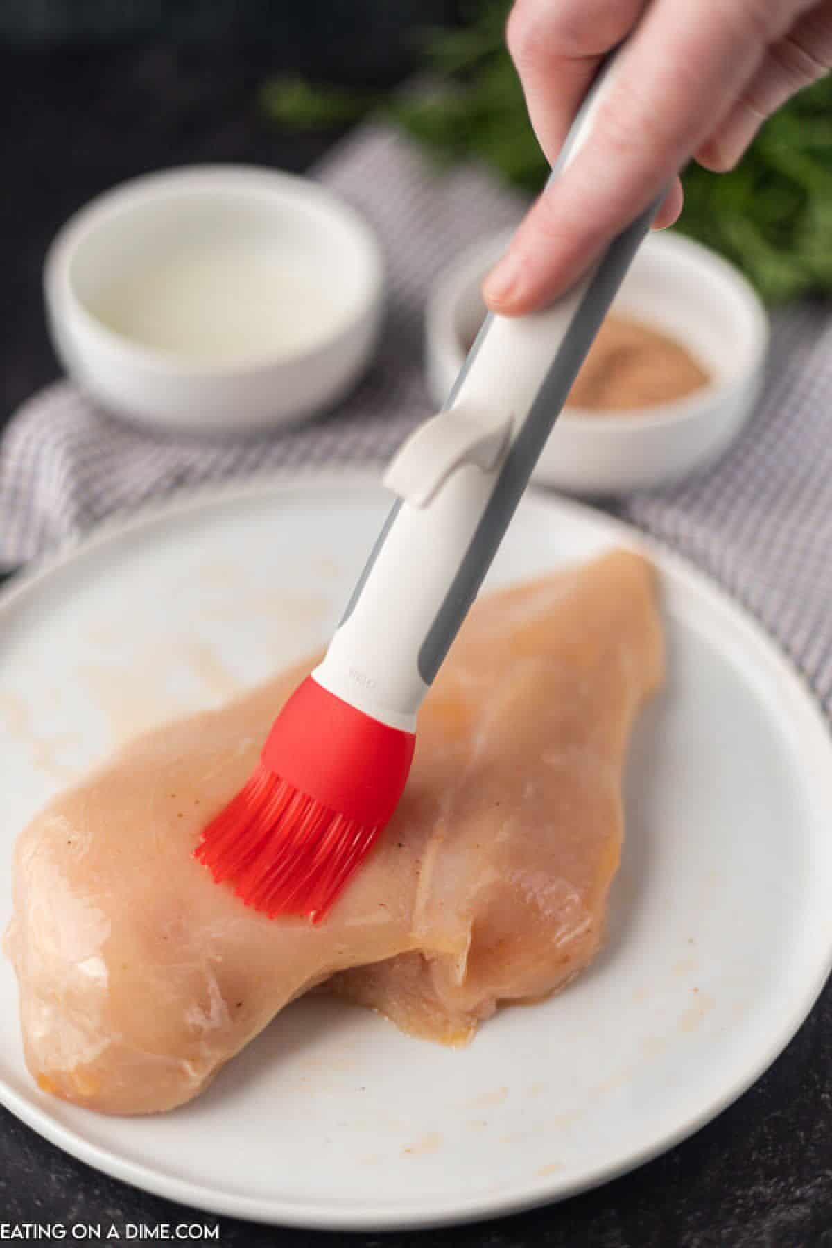 A photo showing the basting brush brushing the oil on a chicken breast on a white plate 