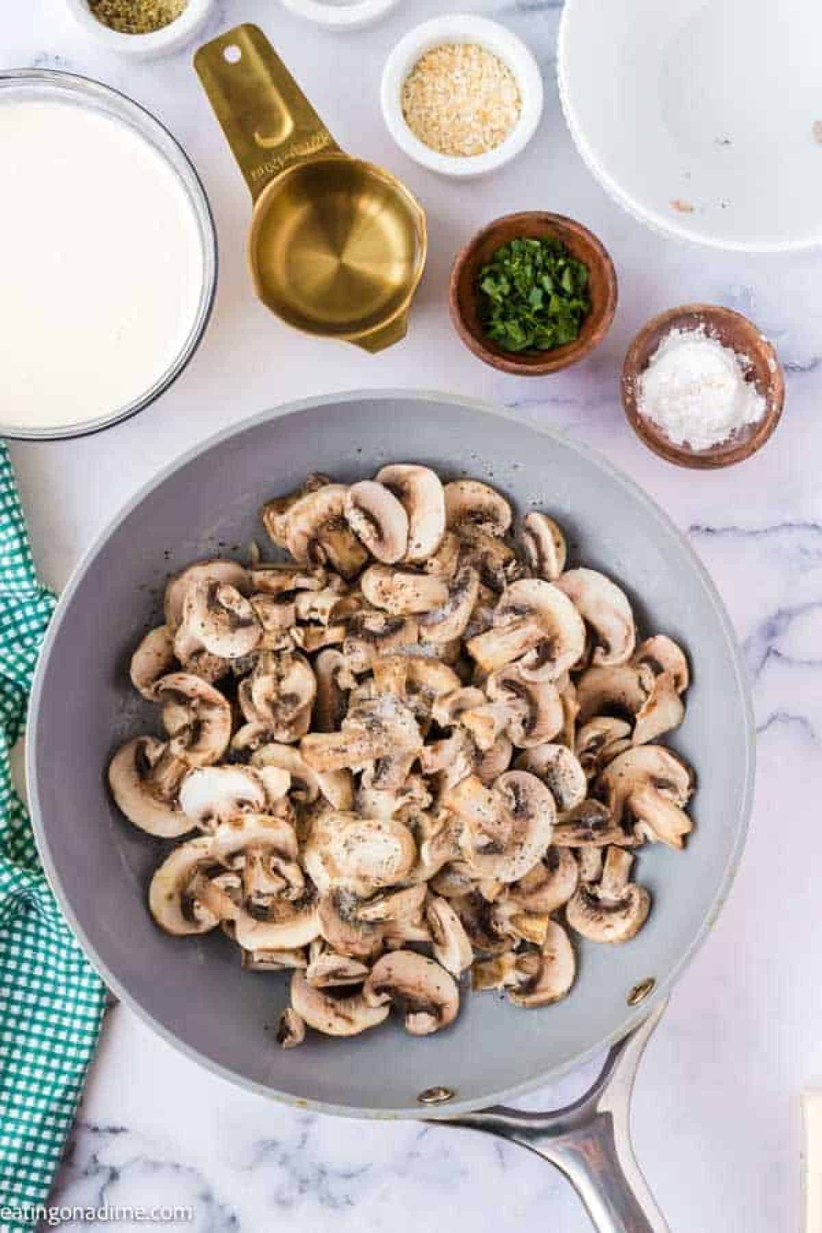 Slice mushrooms in a skillet topped with seasoning with bowls of heavy whipping cream and small bowls of seasoning