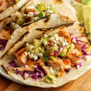 Three grilled fish tacos filled with seasoned fish, shredded purple cabbage, diced tomatoes, avocados, and topped with crumbled cheese sit on a white plate. The tortillas are lightly charred, and a side of lime wedges is visible in the background. Perfect for Meal Plan 14.